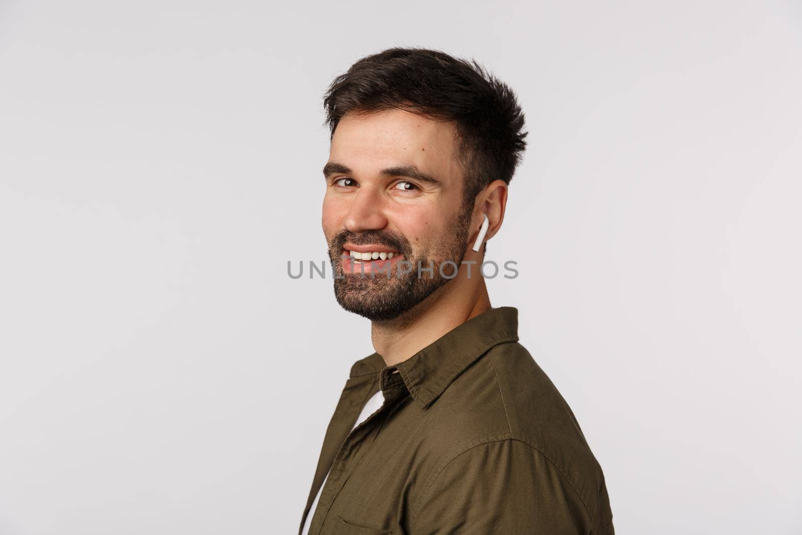 Close-up profile shot cheerful, friendly and happy smiling young man in coat, turn face to camera to laugh and grin pleased, listen music wireless earphones, bought headphones, white background by Benzoix