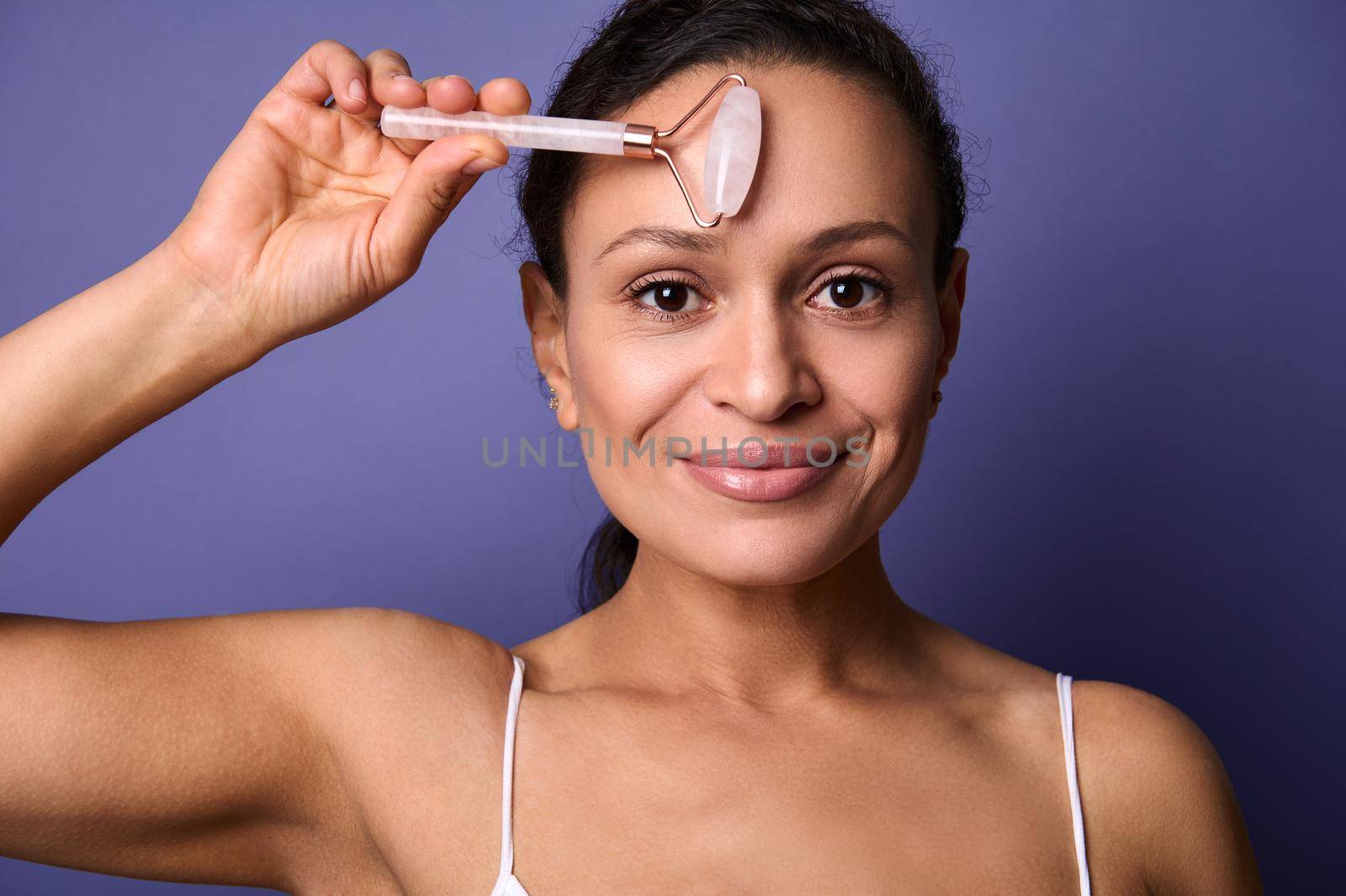 Beautiful middle aged woman with healthy perfect clean shiny skin and natural makeup massaging her face with pink stone jade roller massager, smiles looking at camera, isolated over violet background