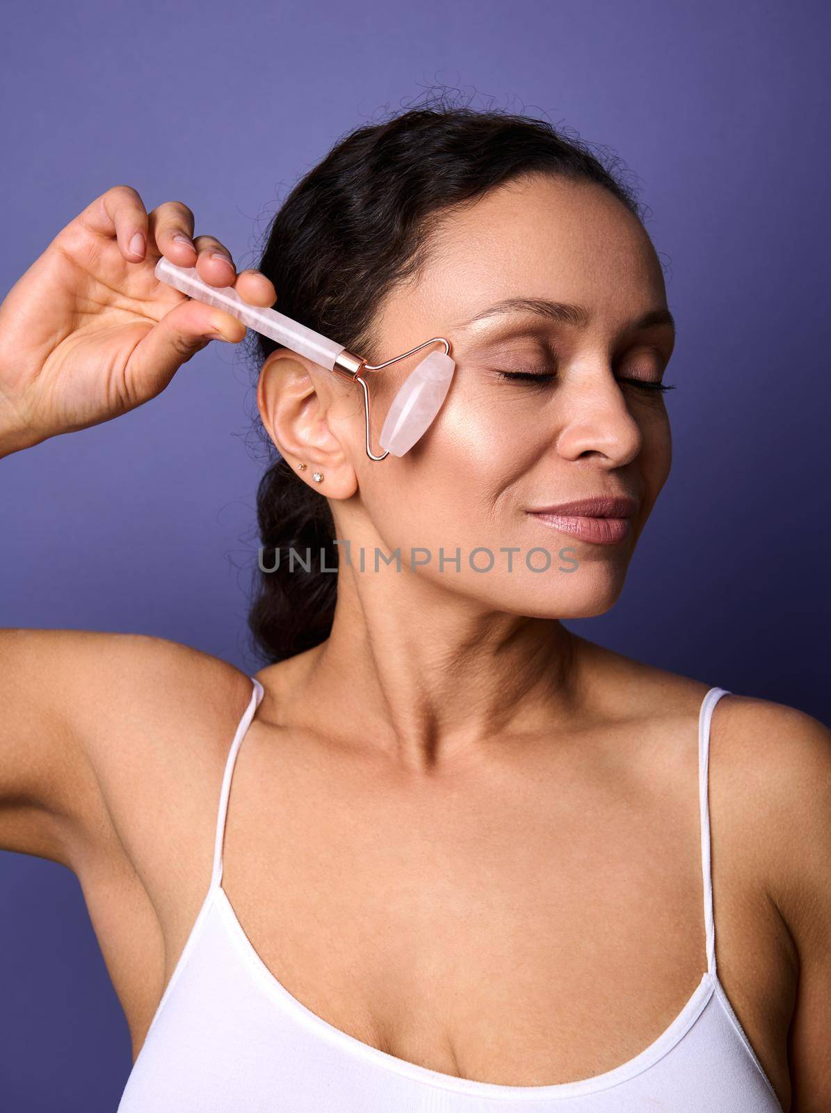 Close-up portrait of dark haired beautiful woman using natural jade stone roller facial massager. Facial massage for skin care, smoothing and anti aging beauty treatment on purple isolated background by artgf