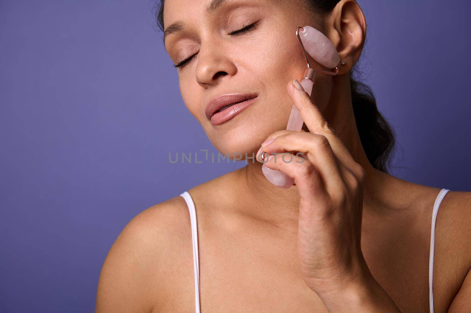 Pretty woman using cosmetics jade stone roller for face massage. Beautiful woman caring of her beauty face with cosmetic tool, performing lymphatic drainage facial lifting massage. Skin care concept