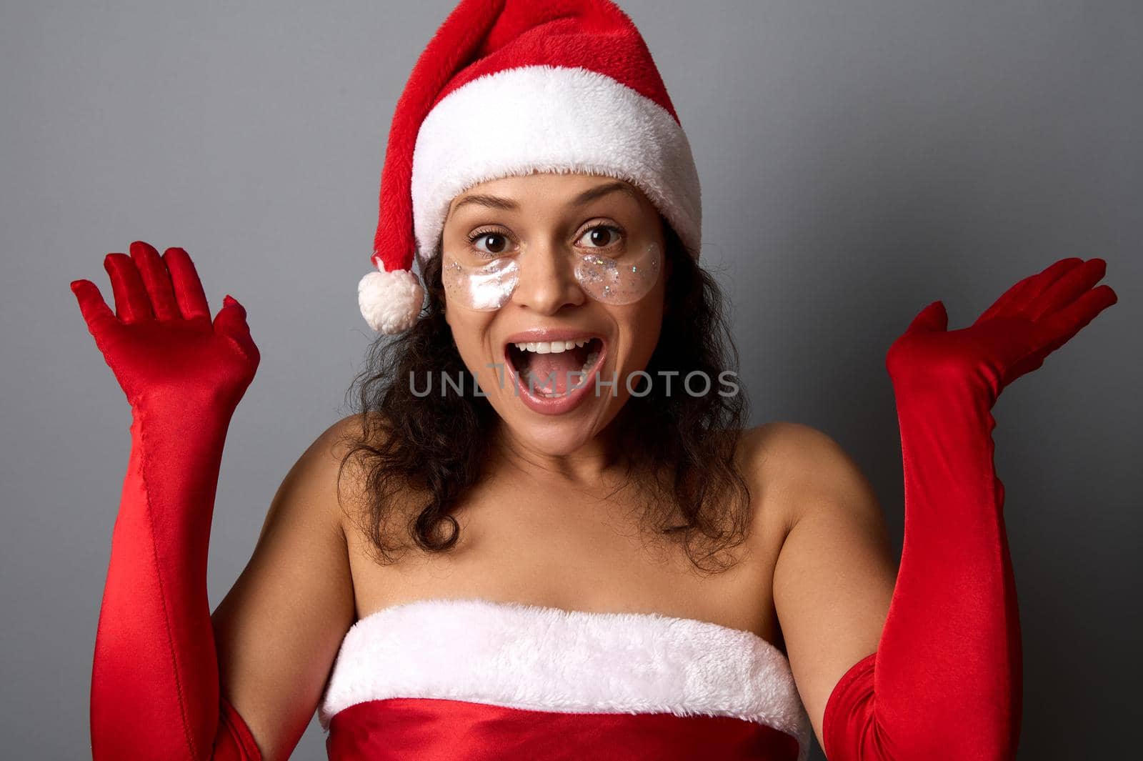 Happy beautiful woman with shiny smoothing eye patches under eyes , dressed in Santa attire puts her hands palms up and rejoices looking at camera. Merry Christmas, New Year and cosmetology concept by artgf