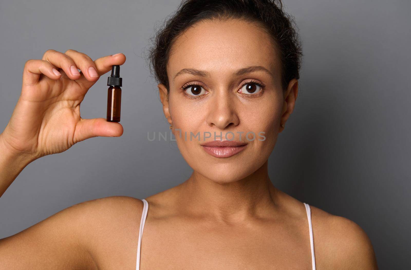 Beauty portrait of woman with perfect fresh clean skin looks at camera posing over gray background holding a bottle with beauty product. SPA, cosmetology, beauty treatment concept with copy ad space by artgf