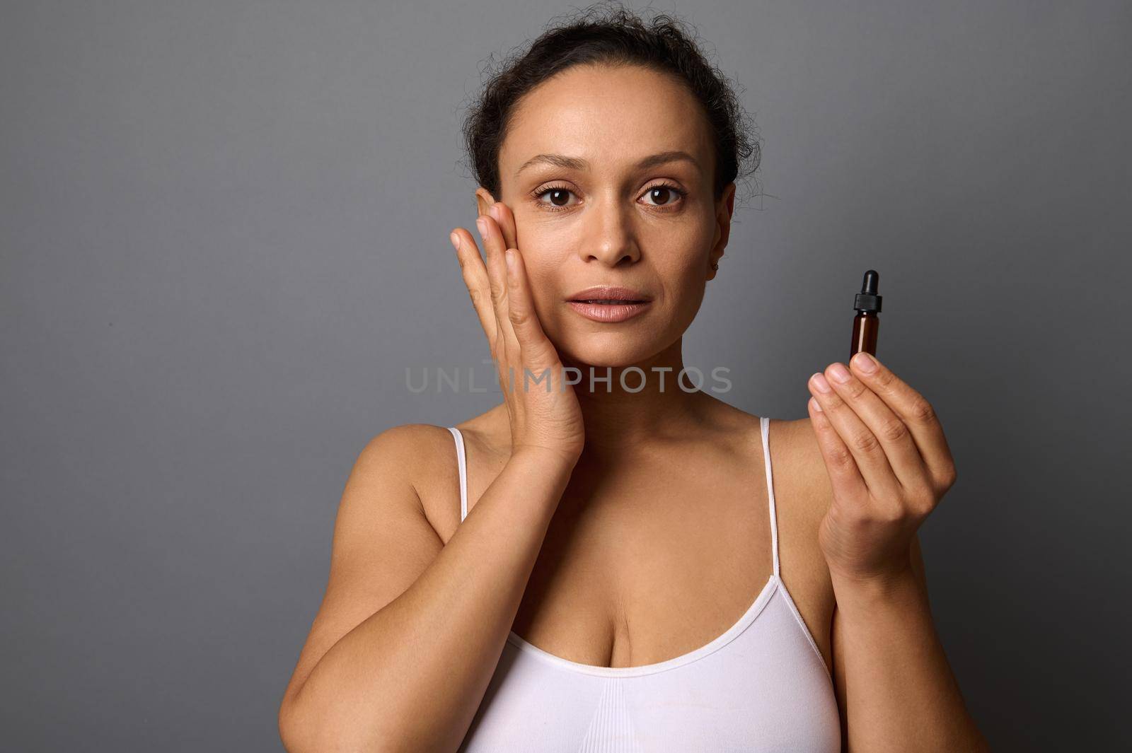 Beautiful middle aged woman holds a jar with beauty product, applies it on her face and performs anti-aging lymhpatic drainage massage for rejuvenating and skin smoothing. SPA, cosmetology concept