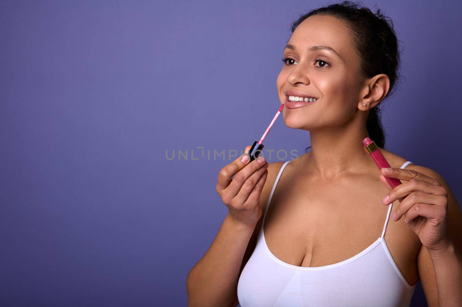 Beautiful African American woman in white underwear applies lipstick, hygienic lip gloss to plump lips, smiles toothy smile, posing over purple background with advertising copy space. Make-up concept by artgf