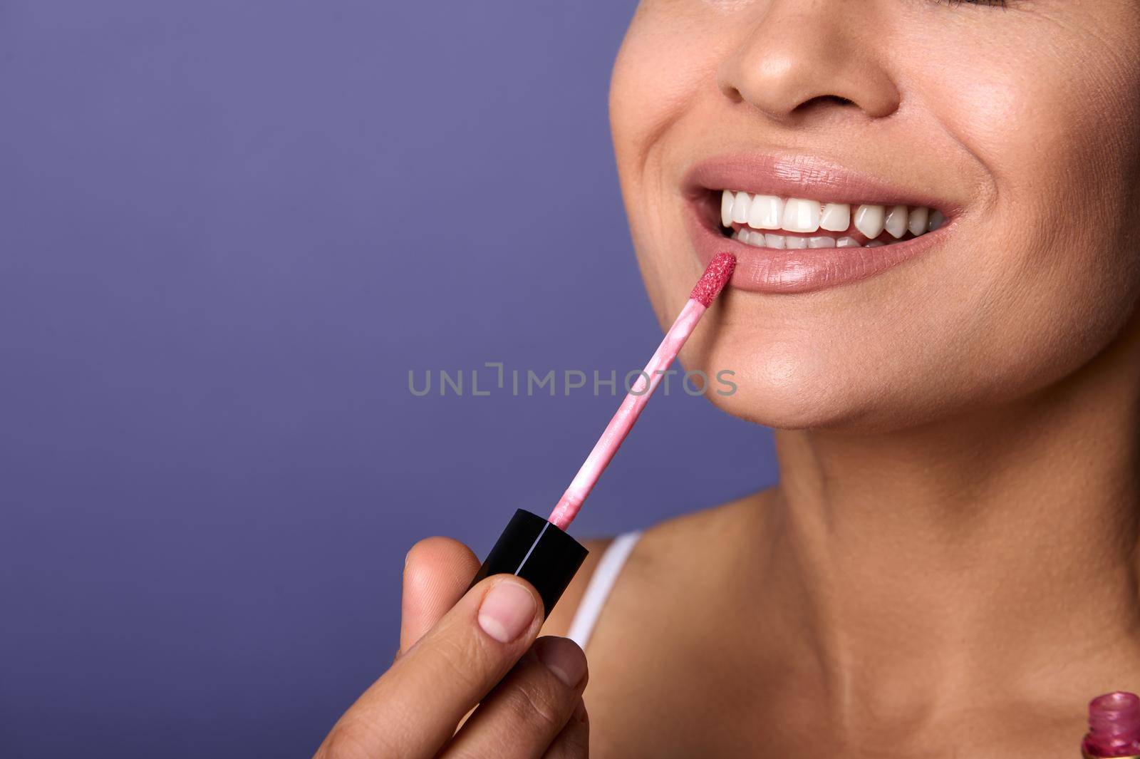 Close-up. Cropped view of attractive smiling woman with perfect clean healthy skin applying lip gloss hygienic balm on plump lips isolated on purple background. Copy ad space. Make-up concept by artgf