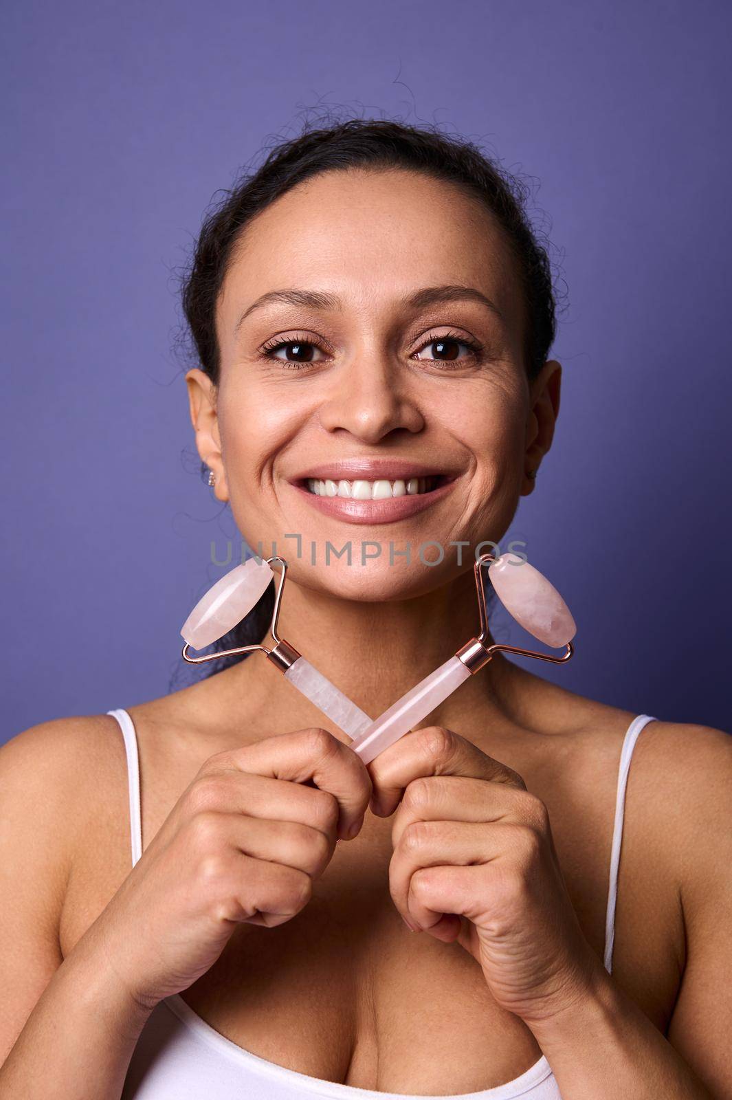 Close-up portrait of a natural beauty middle aged attractive Hispanic woman, smiling with cheerful toothy smile holding two pink jade roller stone massager in her hands, isolated on violet background