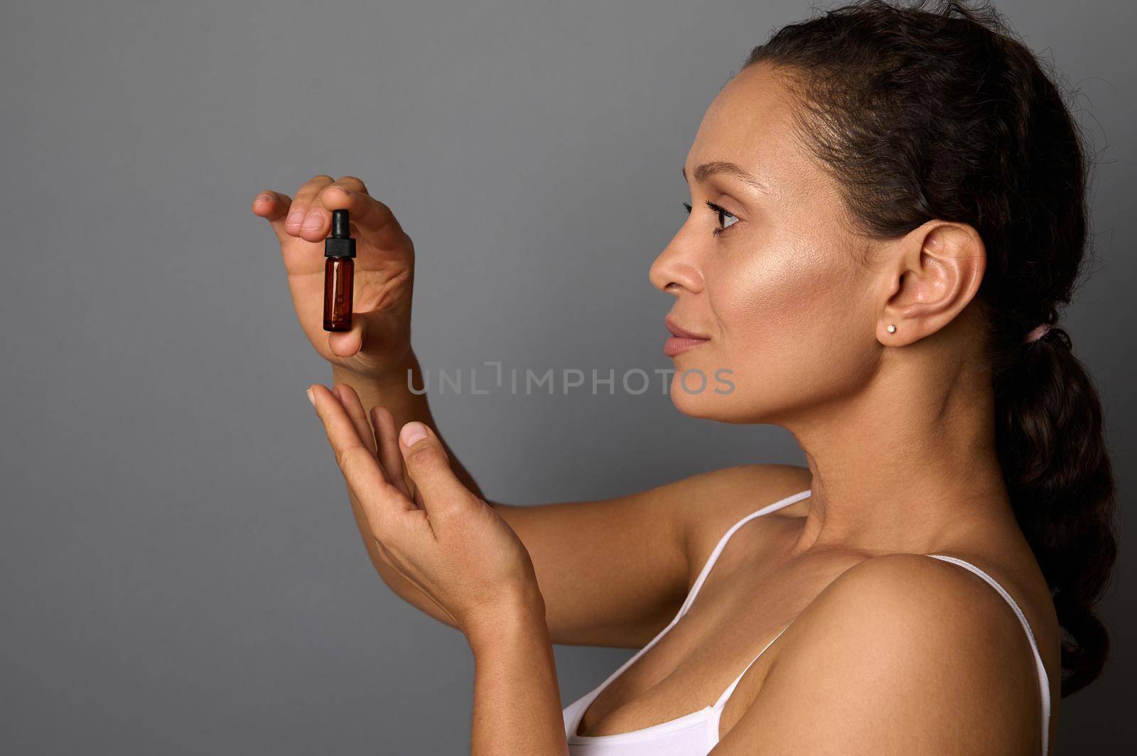 Middle aged African woman holding a dark glass bottle with cosmetic beauty product and dropper, over gray background. Beauty treatments, smoothing, rejuvenation, moisturizing, smoothing concept