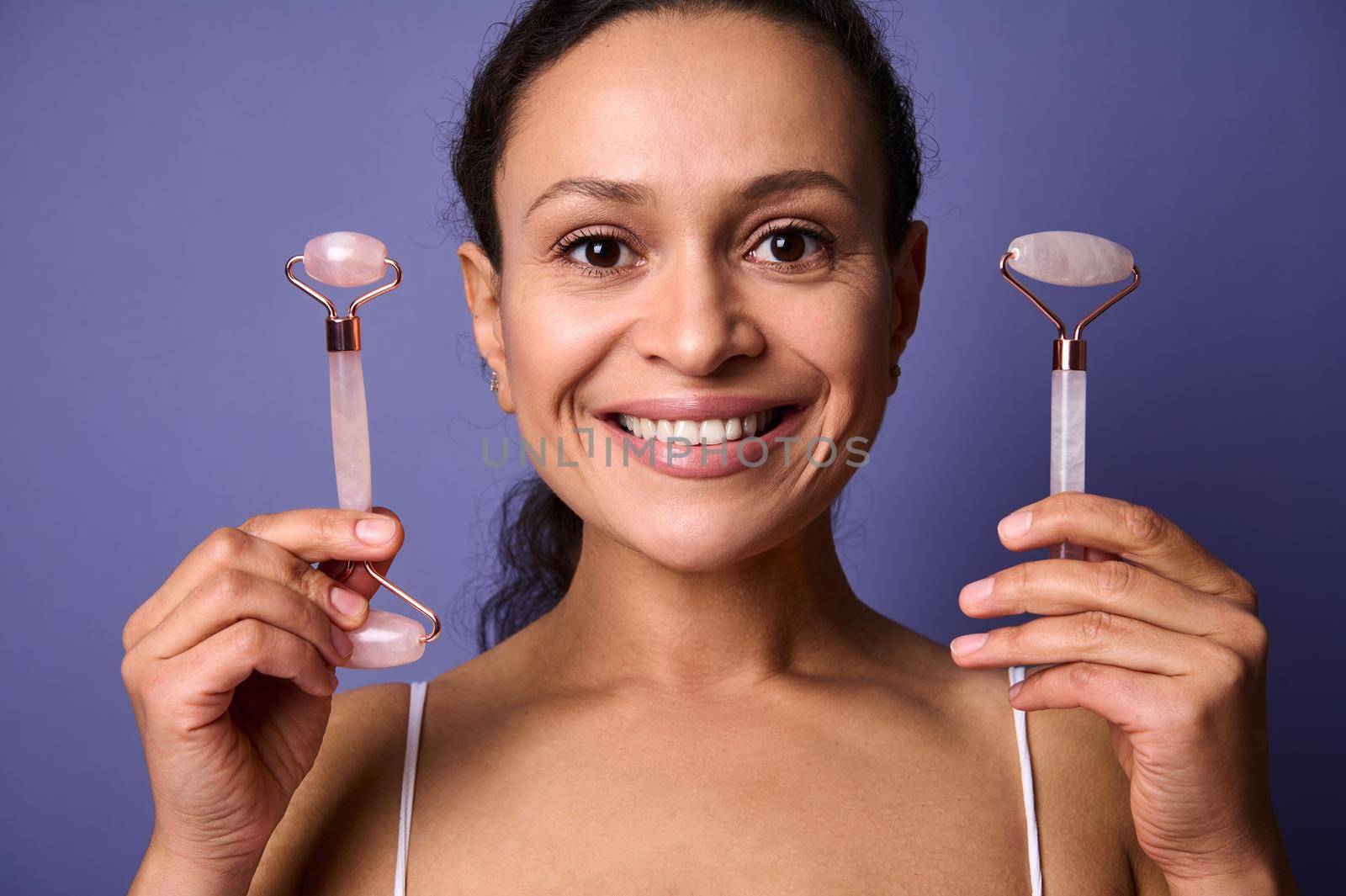 Concept of skin rejuvenation. Face lift anti-aging treatment. Middle aged pretty woman smiles toothy smile showing her face posing with jade stone roller massage on violet background with copy space