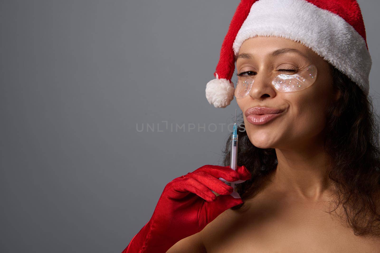 Sexy brunette in Santa hat, wears under eyes patches, attractively looks at camera, holds syringe with beauty injection against gray background. Copy space for Christmas ad. Anti-aging cosmetology by artgf