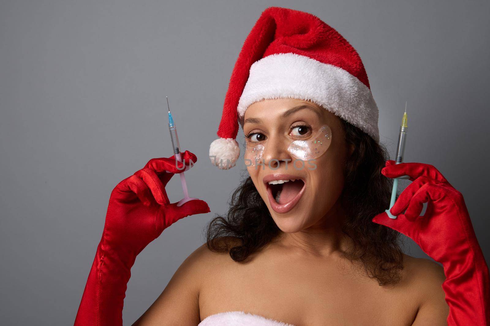 Sexy pretty woman in Santa hat, wears under eyes patches, rejoices looking at camera, holds syringes with beauty injection against gray background. Copy space for Christmas ad. Anti-aging cosmetology by artgf