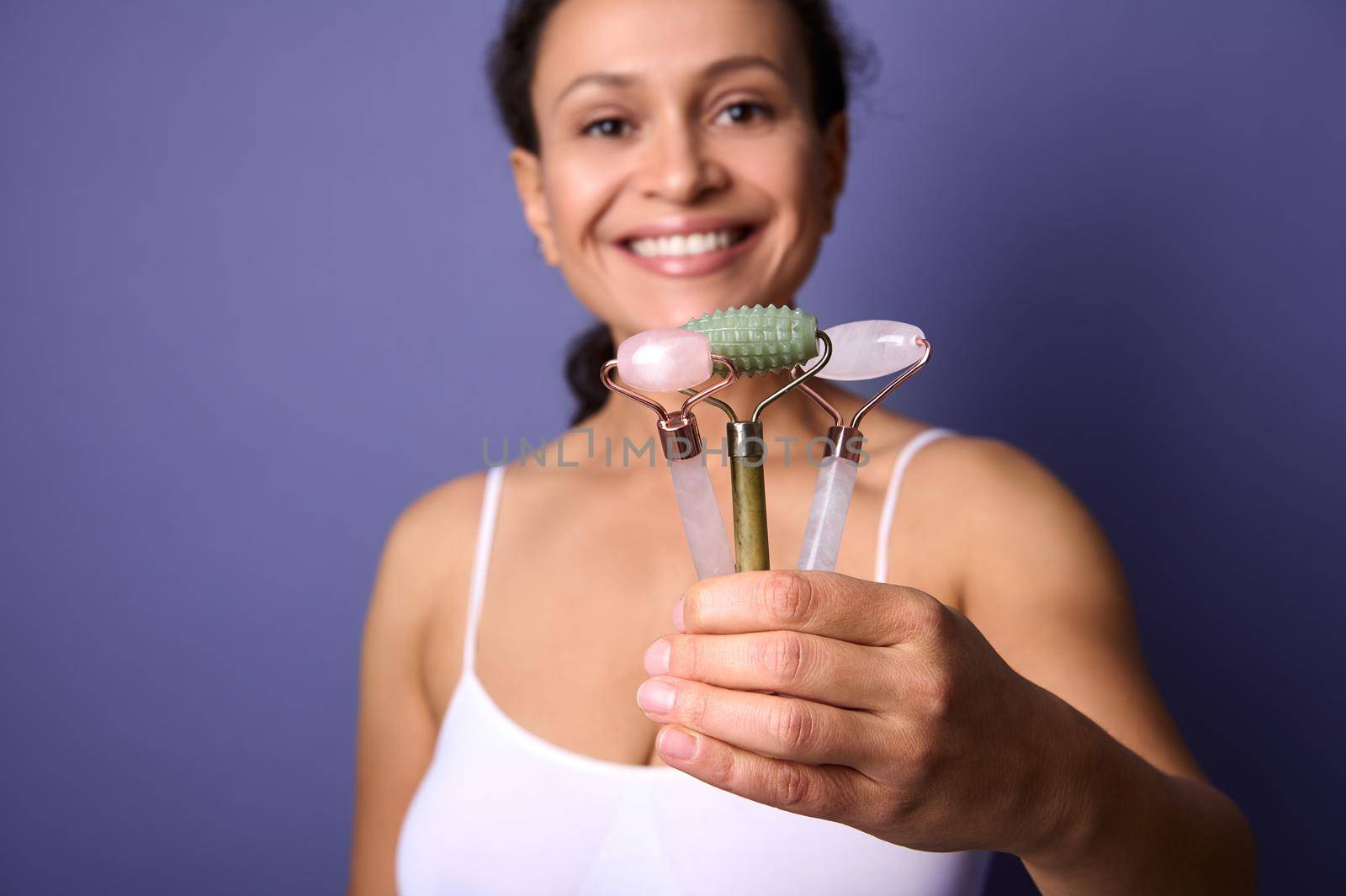 Focus on colorful Ayurvedic jade stone roller massagers in the hand of cheerful smiling middle aged pretty woman posing against violet background. Face and skin care concept with copy space. Close-up by artgf