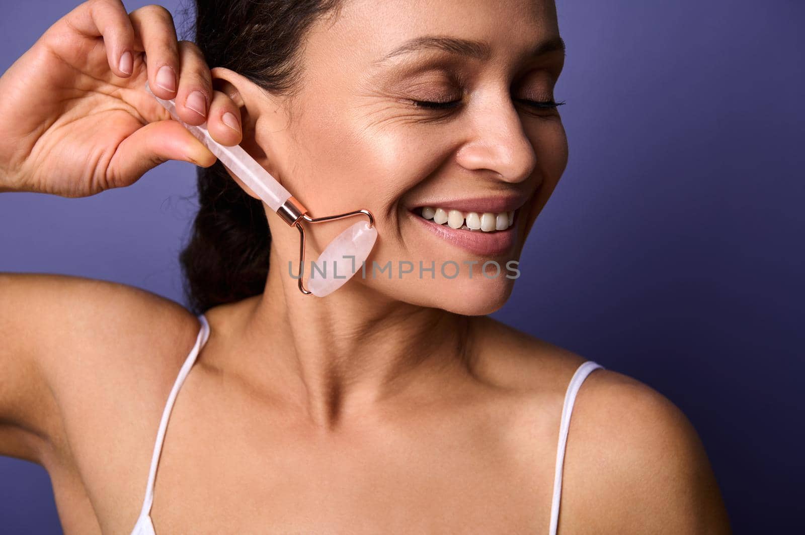 Close-up portrait of smiling African pretty woman doing face lifting smoothing anti-aging massage with pink jade stone roller, isolated over violet background with copy space. Face skin care concept