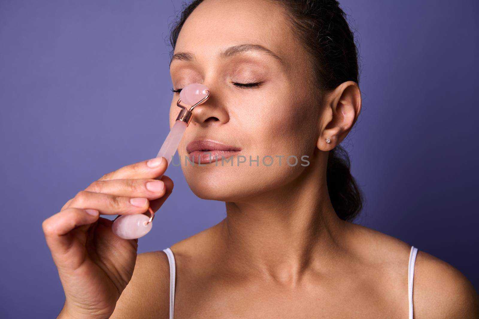 Close-up beauty portrait of middle aged pretty woman massaging her nose with stone roller massager, doing smoothing anti-aging face lifting massage with cosmetic tool, isolated on violet background