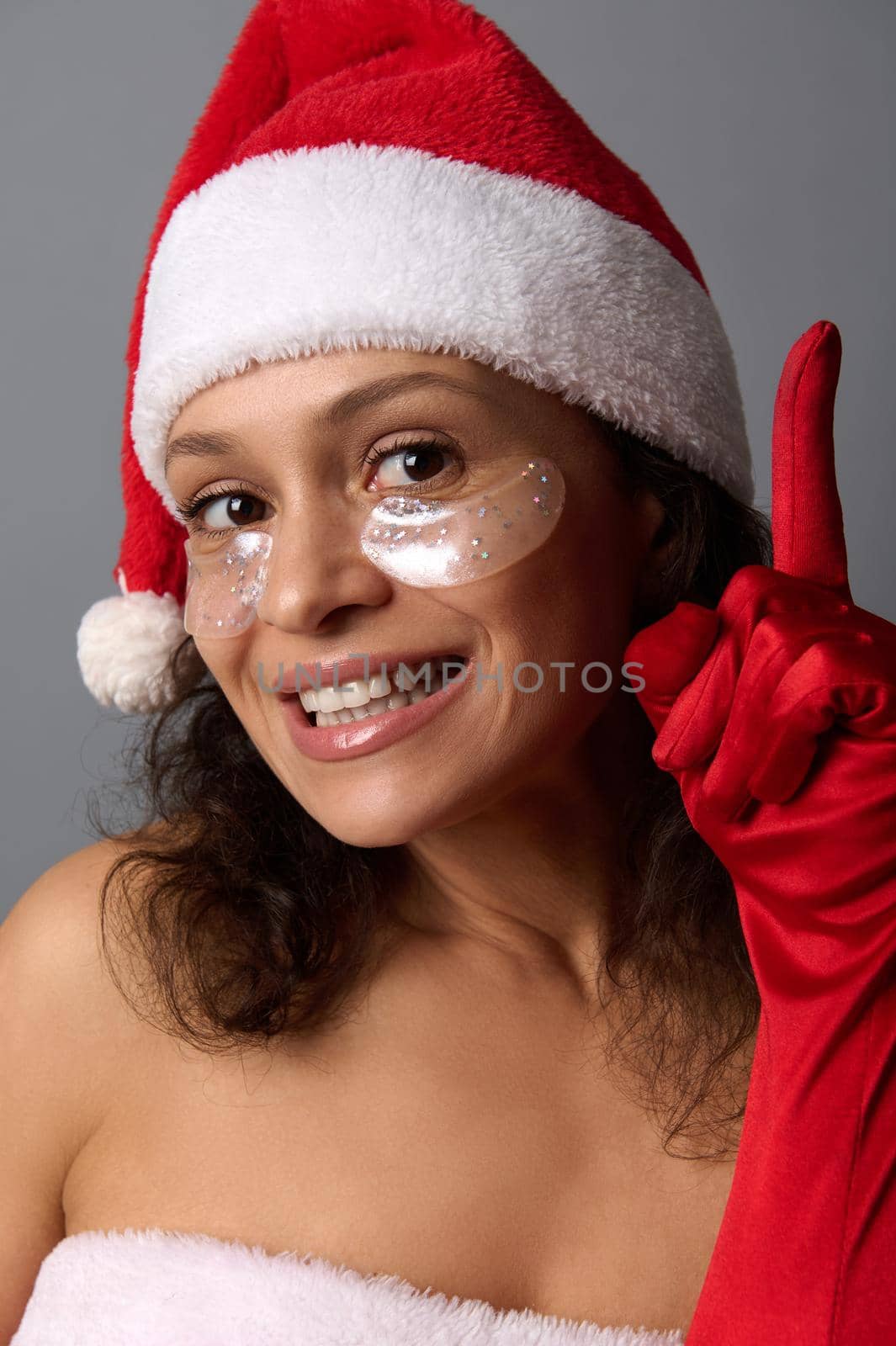 Close-up beauty face portrait of attractive young brunette with beautiful smile, wearing Santa hat and smoothing patches under eyes points finger up on copy space on gray background, looking at camera by artgf