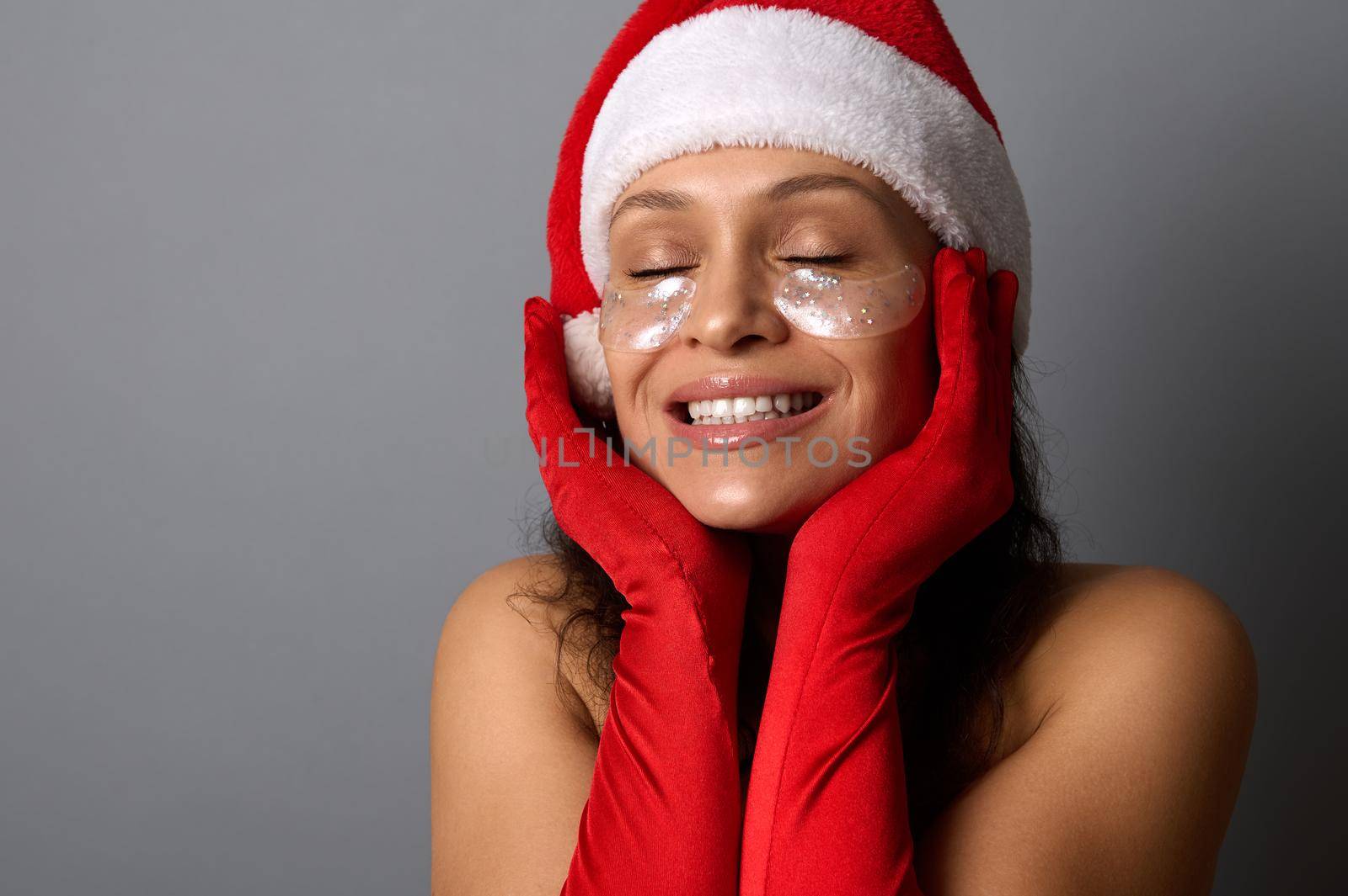 Young woman in Santa carnival outfit, with shiny smoothing spots under her eyes, holds her hands near her face, smiles with a beautiful toothy smile and poses with closed eyes on a gray background by artgf