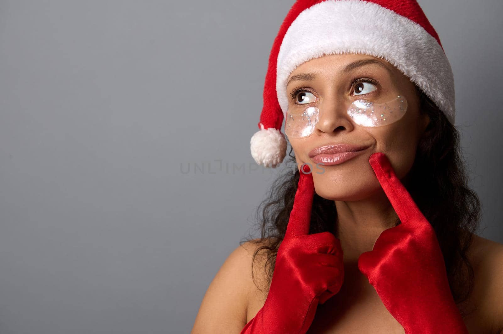 Pretty woman with smoothing eye patches , wearing Santa Claus hat and red satin gloves, presses her fingers to her lips and smiles, looking at copy space on gray background. Spa, skin care concept