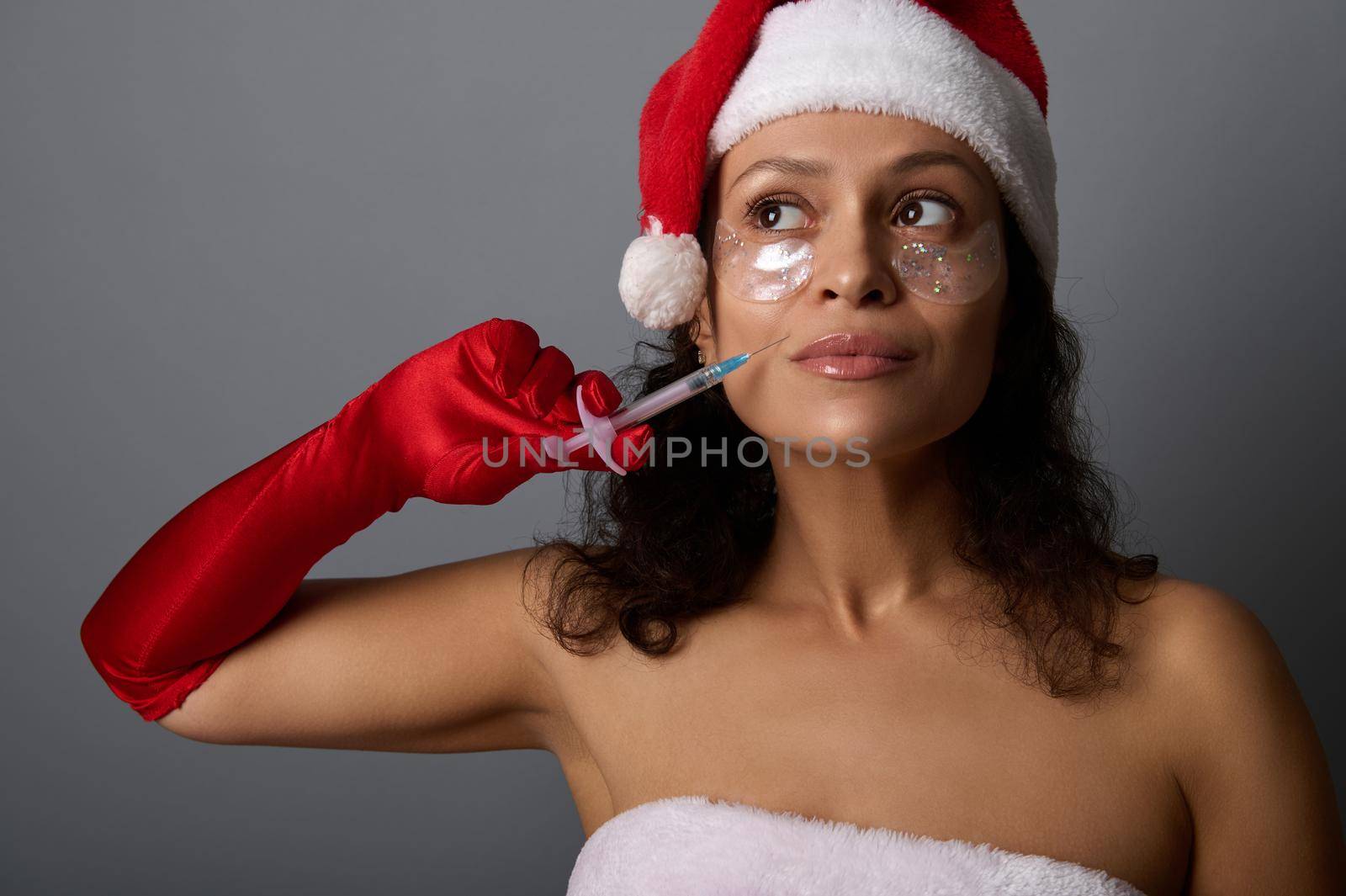 Attractive pretty woman in Santa carnival attire holds syringe on her nasolabial fold and looks up on copy space for advertising for Christmas giveaways for spa salons. Injection cosmetology concept