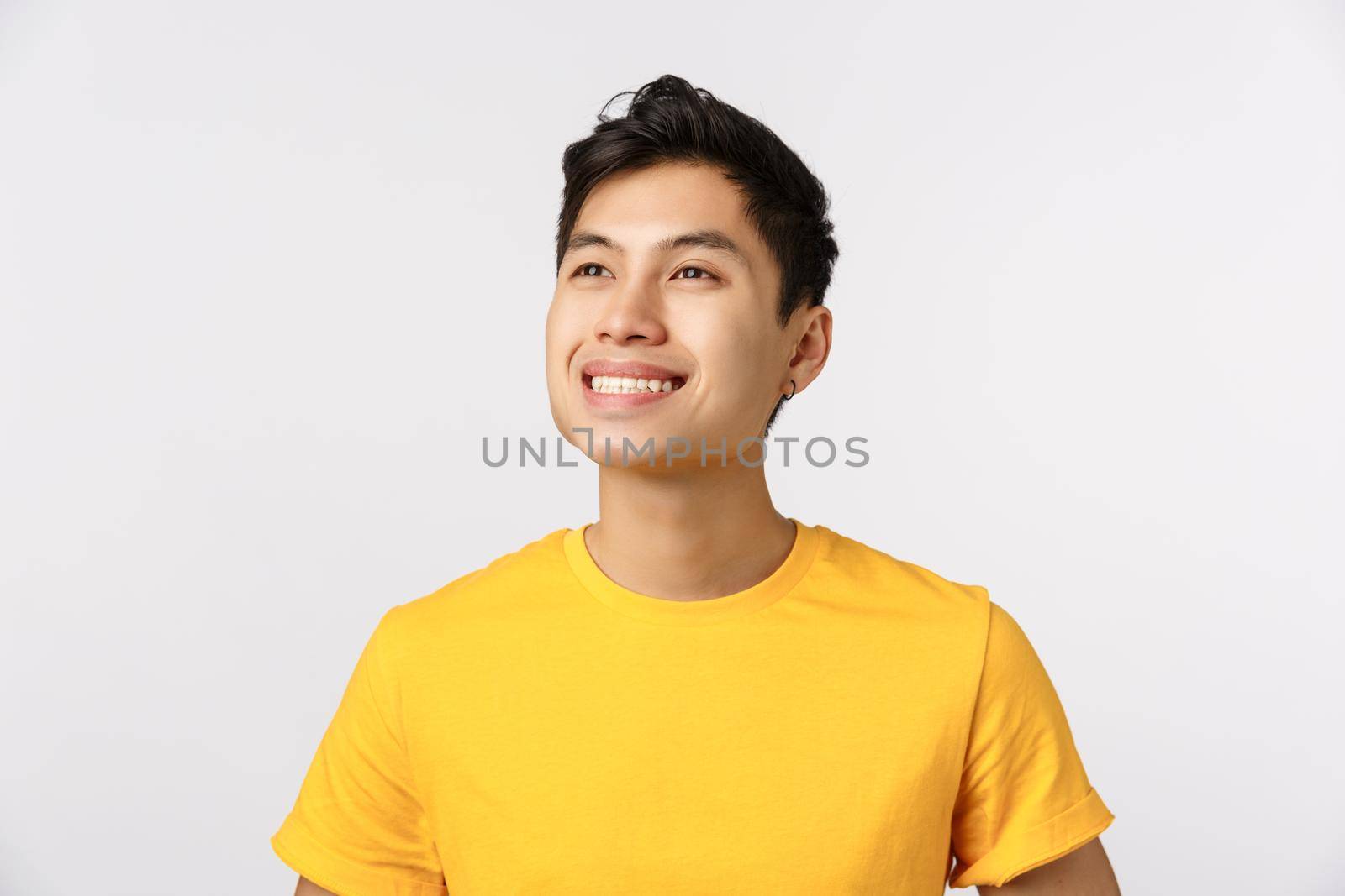 Proud and dreamy good-looking confident and cheerful asian man, wear yellow t-shirt, smiling delighted and contemplate something beautiful at upper left corner, white background. Copy space