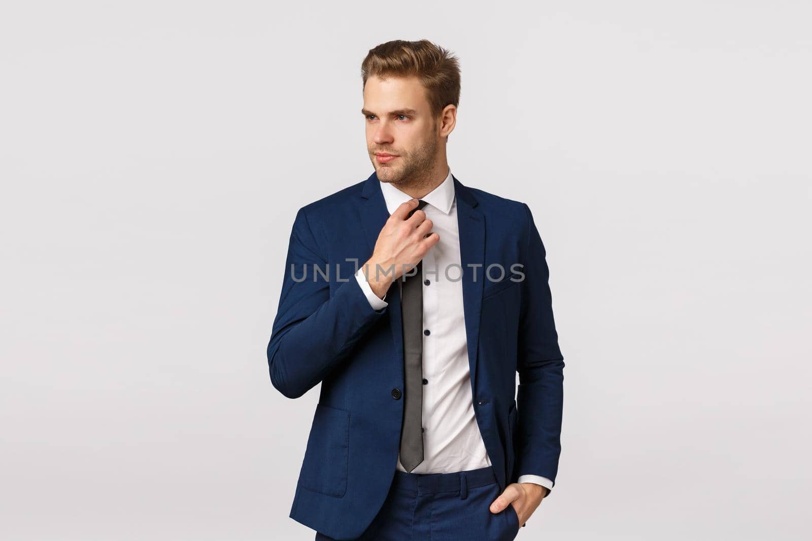 Serious-looking sassy businessman in blue classis suit, adjusting tie and looking away, holding hand in pocket, getting ready for work, waiting cab in downtown to go business meeting, greet partners.