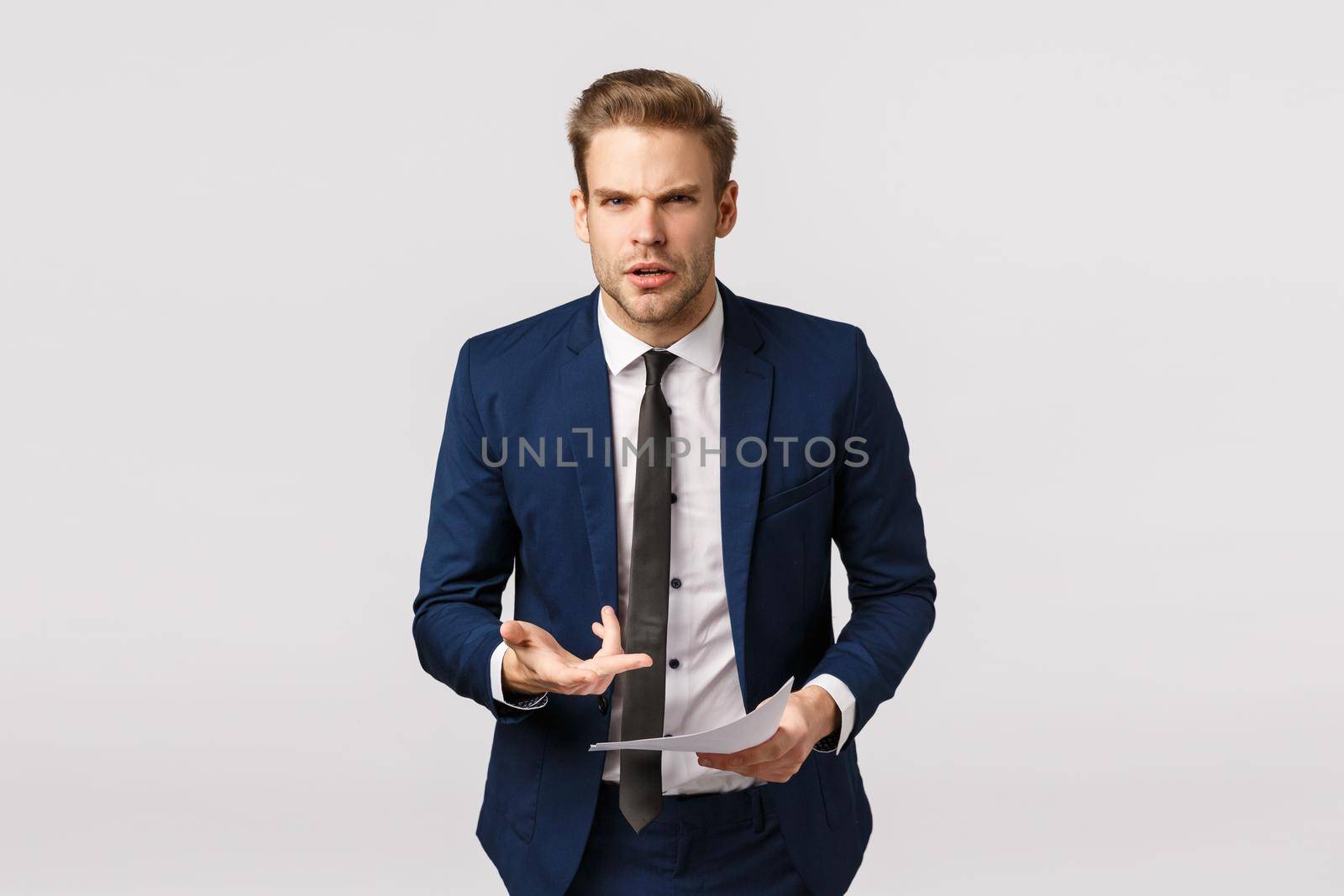 Furious, aggressive young businessman in classic suit, frowning staring outraged and pointing at documents pissed, scolding employee writing bad report, standing bothered white background.