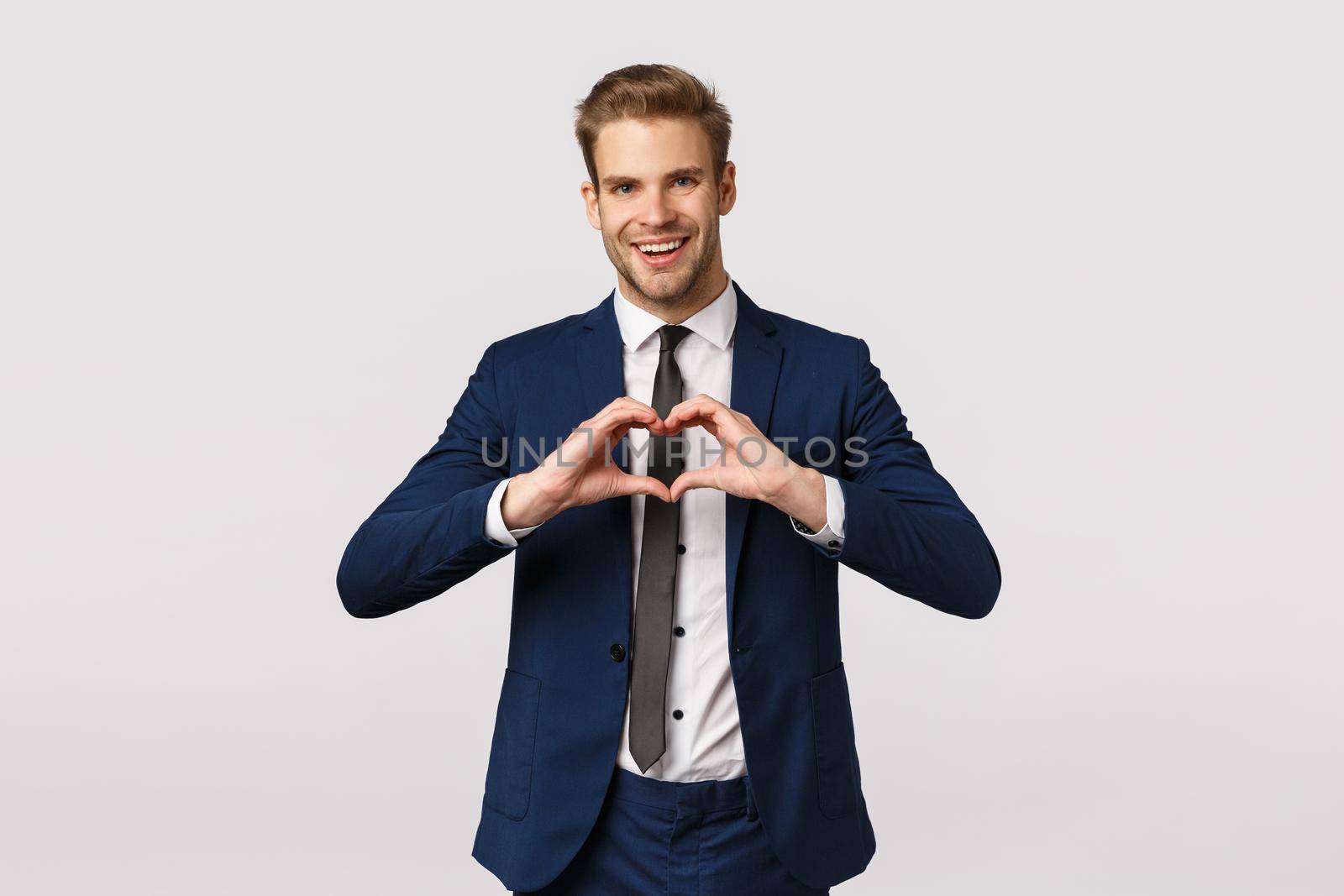 Joyful bearded young blond man in blue classic suit, showing heart sign and smiling, come from office home, make surprise dinner for girlfriend, congratulate with valentines day, white background.
