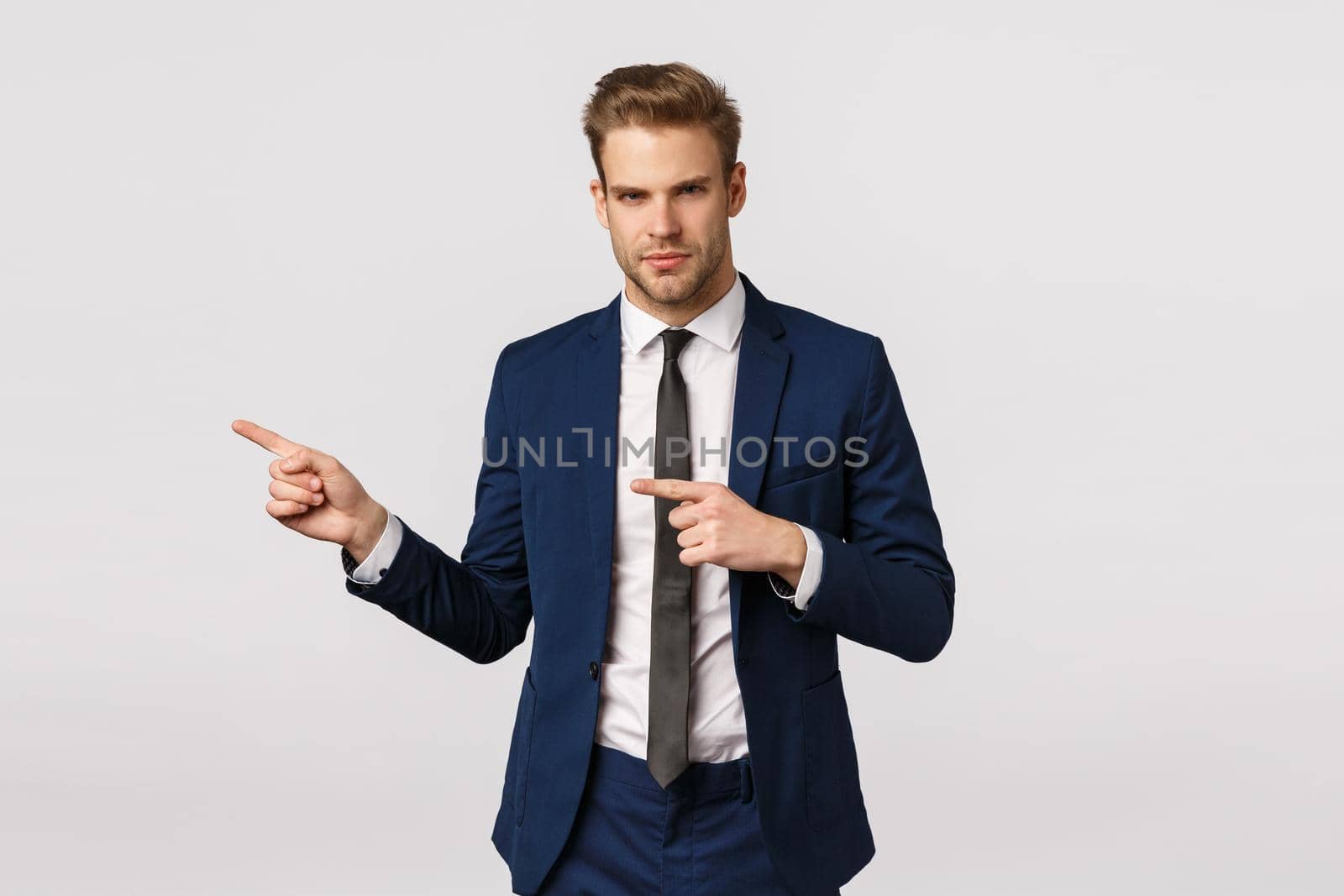 Serious-looking displeased, offended young blond businessman feeling sad, sulking, make grumpy face and looking from under forehead bothered, pointing left, envy someone, white background.