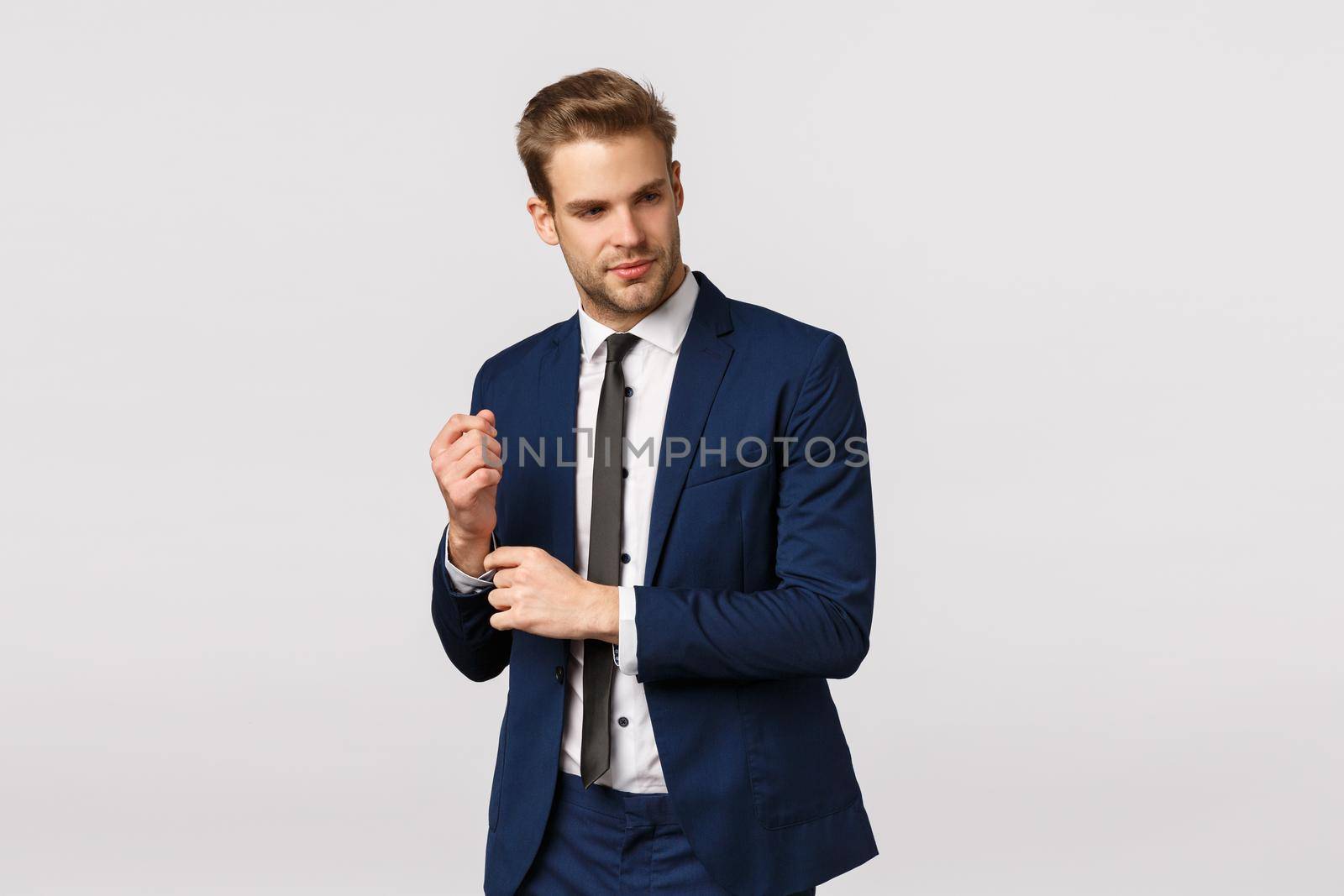 Classy good-looking blond bearded young male entrepreneur in suit, adjusting sleeves of jacket and looking away with pleased smile, relish good deal, knows how achieve success and money.