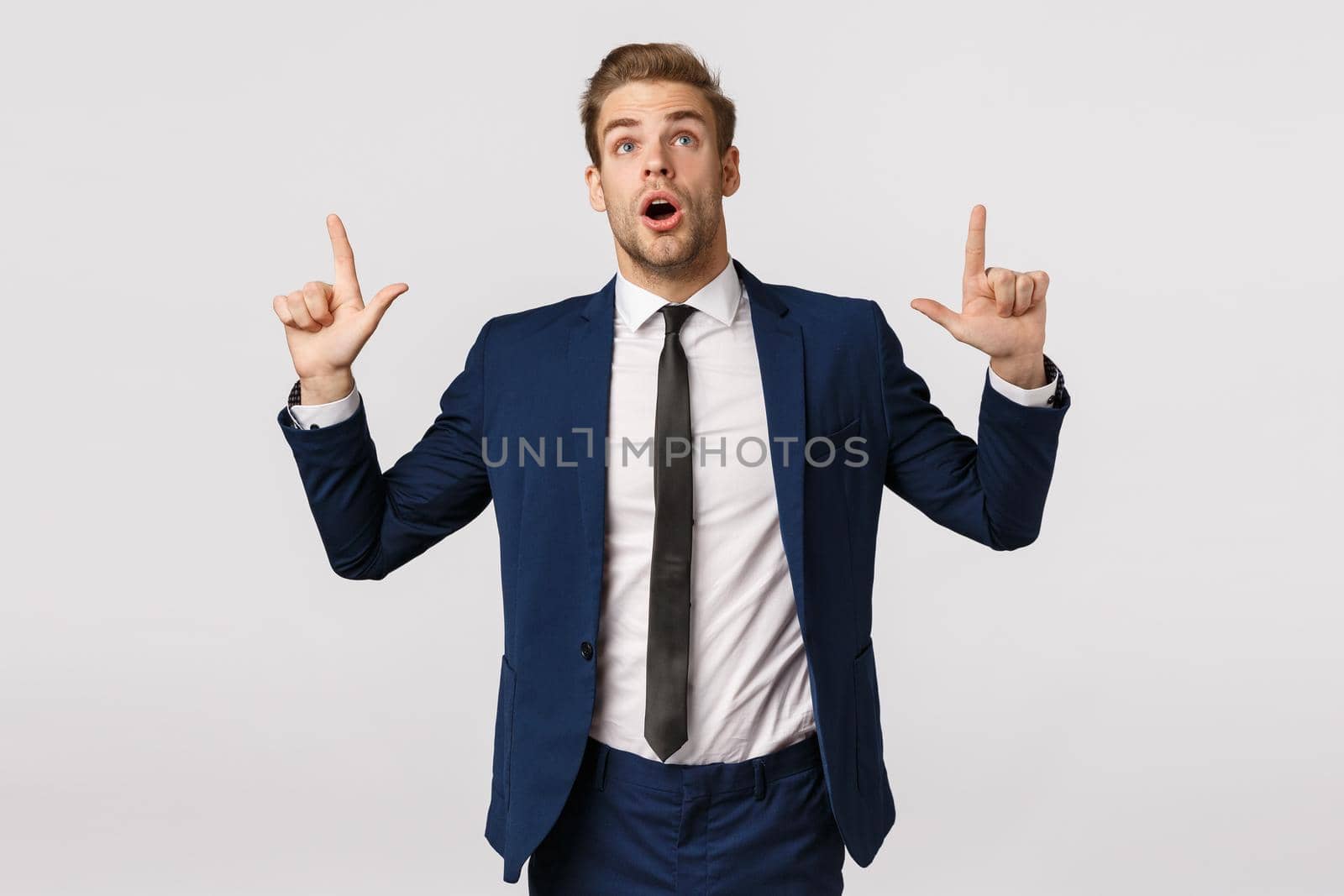 Speechless young man in suit seeing incredible offer, breathtaking scene. Attractive young businessman unshaved standing white background, looking pointing up, open mouth in awe and amazement.