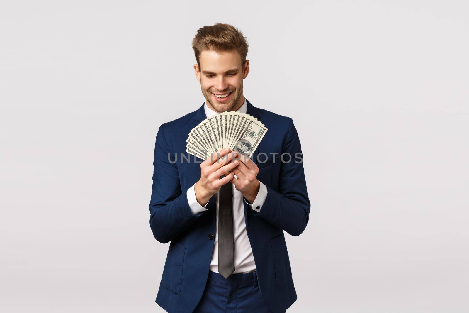 Happy and excited, good-looking businessman in classic suit, counting money, holding big cash dollars and smiling with pleased, delighted expression, standing white background glad.