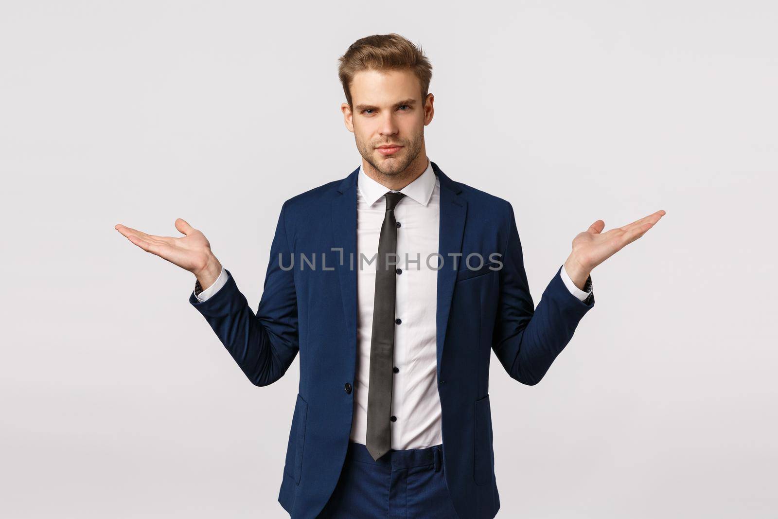 Make your choice. Serious-looking assertive and stylish young blond businessman in classic suit, propose two variants gain money, become rich, spread hands sideways, hold product, white background by Benzoix