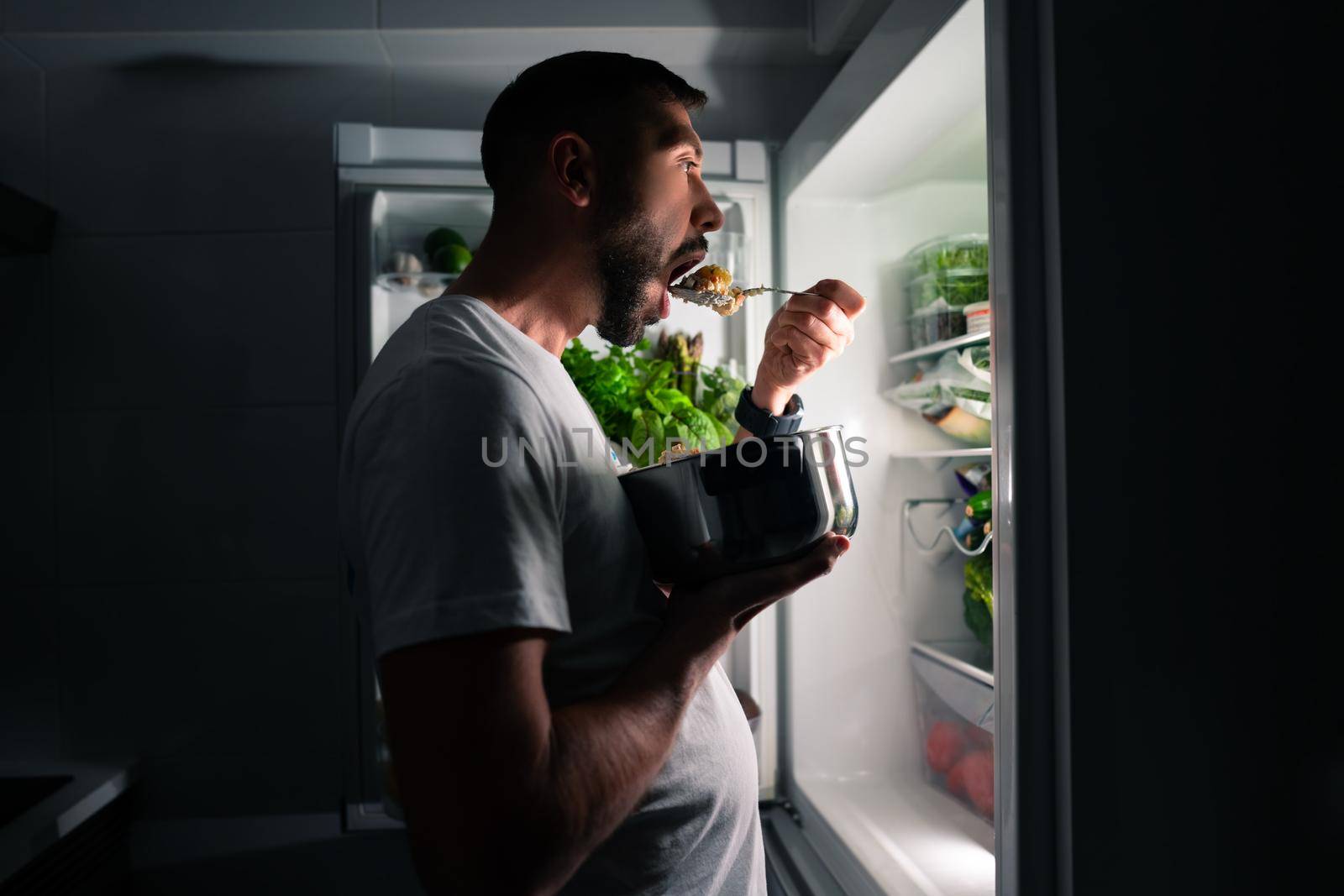 Young hungry man eating food at night and looking in open fridge. Man taking midnight snack from refrigerator by DariaKulkova