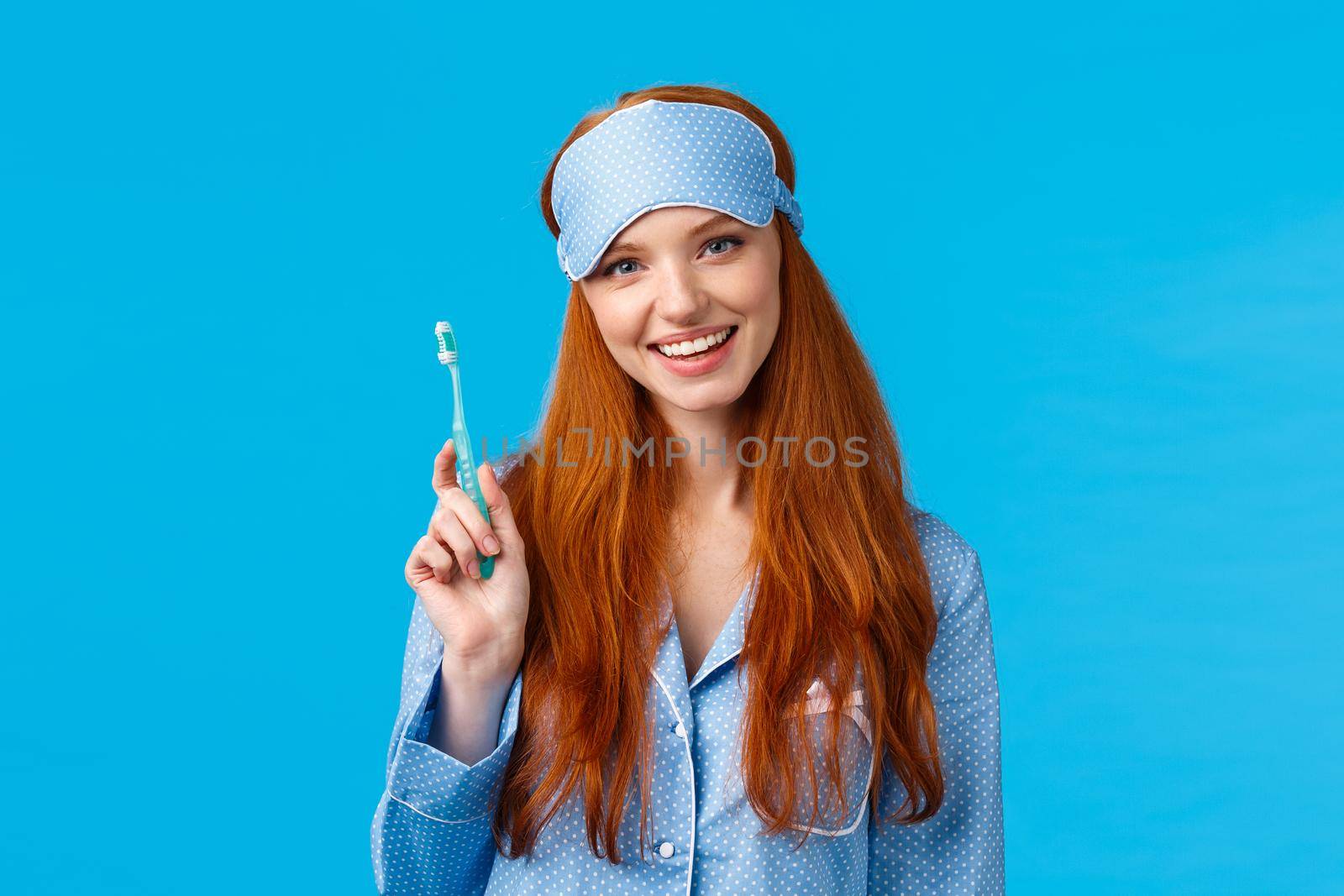 Health, beauty and lifestyle concept. Cheerful pretty european woman with blue eyes, toothy smile, holding toothbrush and smiling delighted, wearing sleep mask, nightwear, blue background.