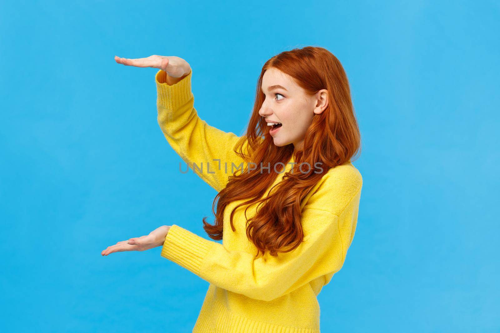 Impressed and startled excited cute redhead female receive big gift, shaping large object or product over copy space, holding hands as if carry enourmous present, smiling astonished, blue background by Benzoix