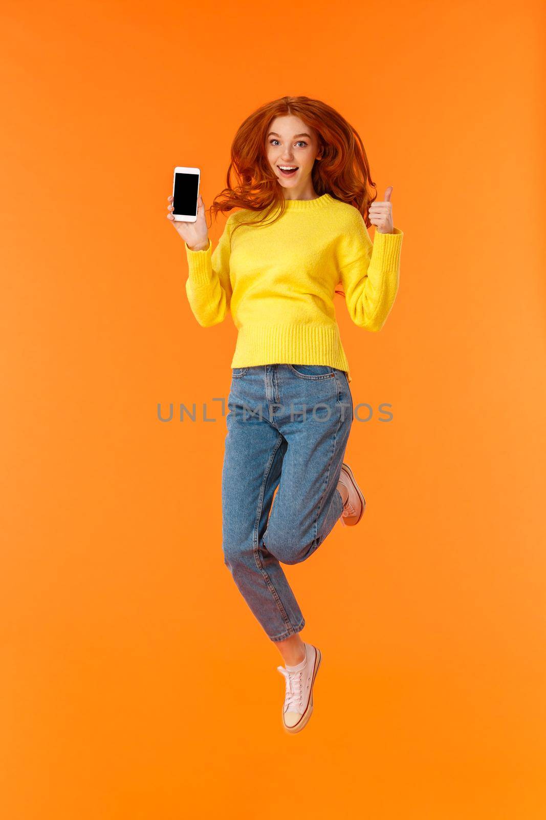 Advertising, technology and holidays concept. Vertical full-length shot cheerful gorgeous ginger girl, redhead woman jumping over orange background with smartphone, show thumb-up by Benzoix