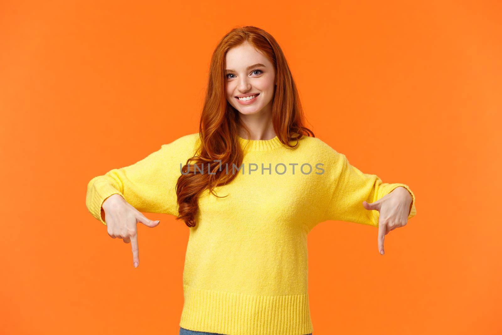 You need see this, check it out. Cheerful gorgeous redhead girl in yellow sweater, smiling and pointing down, recommend product, advertising shopping holidays sales, orange background.