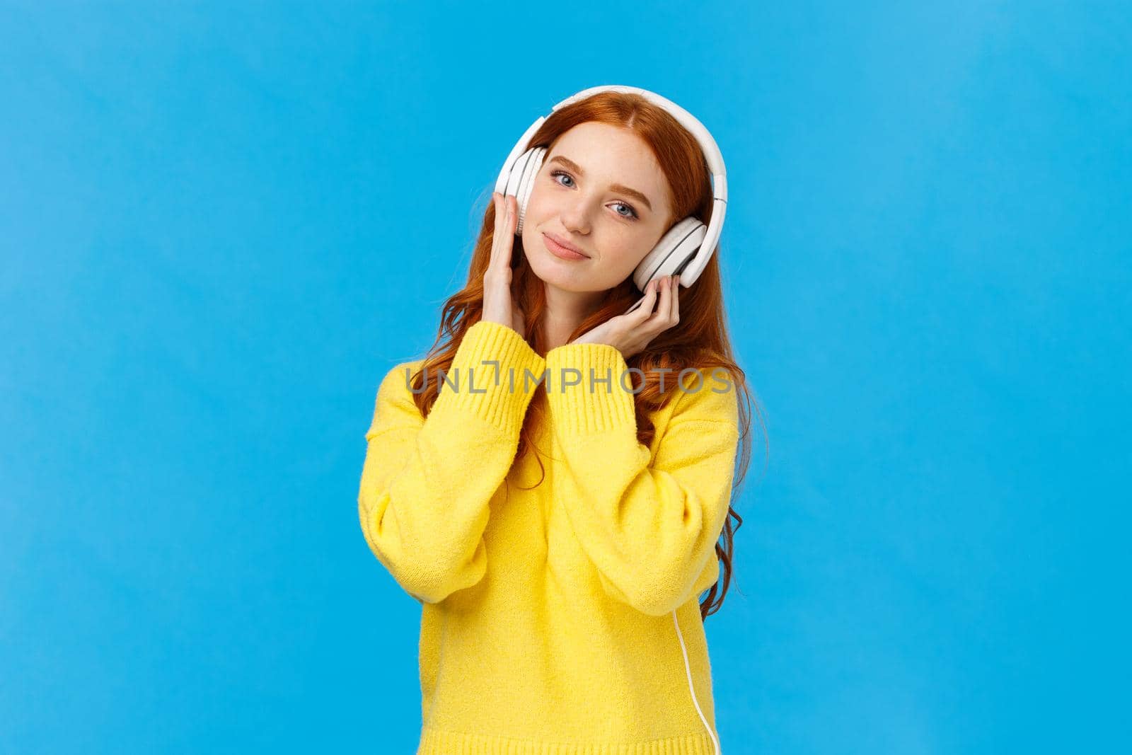 Waist-up shot tender and cute, lovely redhread woman in yellow sweater, tilt head, wear headphones, touching earphones as press to ears, listen music, smiling camera delighted, blue background.
