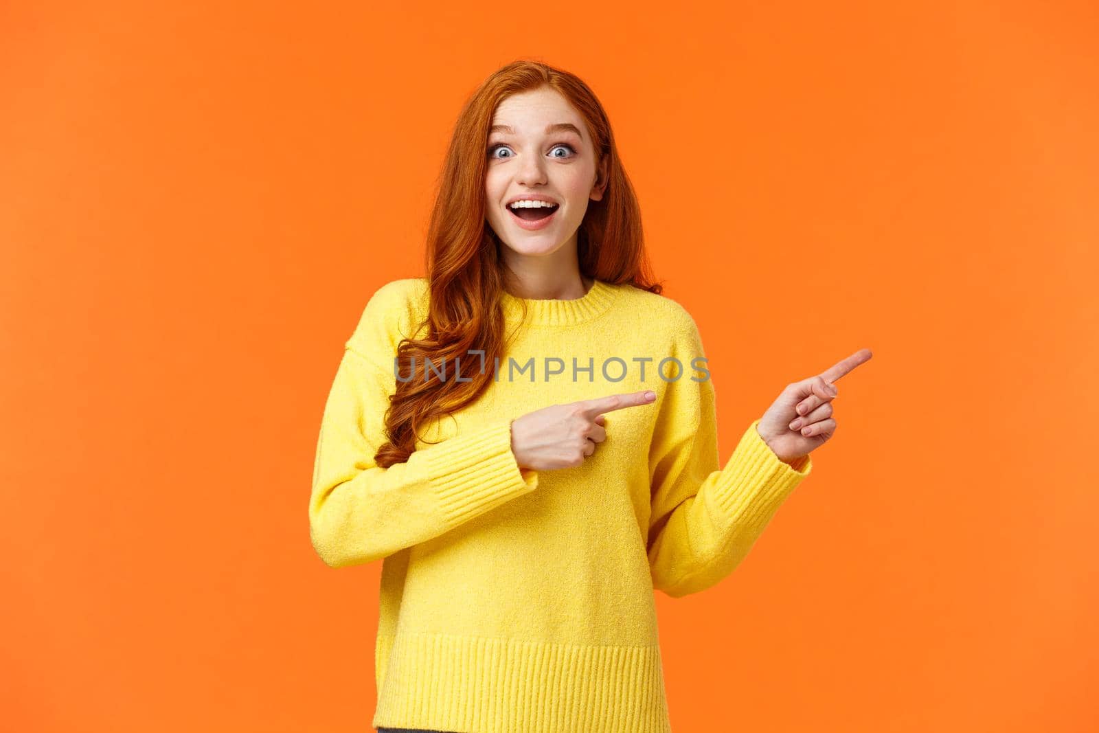 Excited, surprised and fascinated good-looking redhead hipster girl in yellow sweater, got amazing gift for winter holidays, showing people, pointing right and stare camera astounded by Benzoix