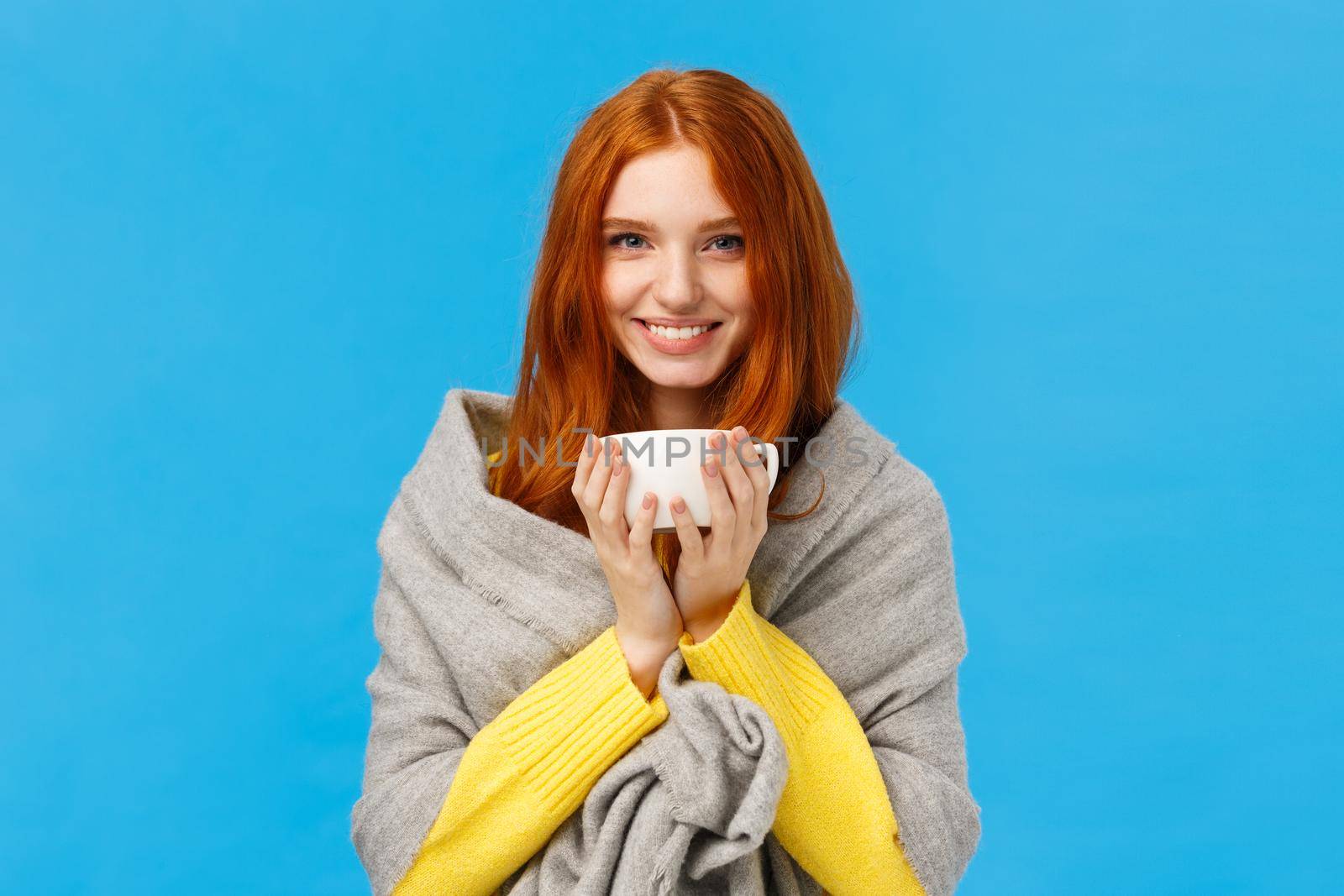 Winter holidays, people and emotions concept. Cheerful lovely and romantic redhead woman came home, warming with hot tea and smiling, wrap herself with grey scarf, standing blue background by Benzoix