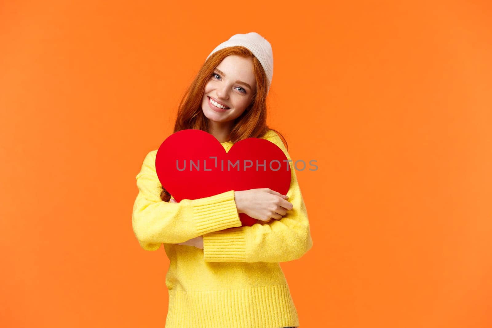 Tender and romantic dreamy redhead woman in winter hat, sweater, embrace big cute red heart sign as symbol of love, smiling joyfully, celebrating valentines day, orange background.