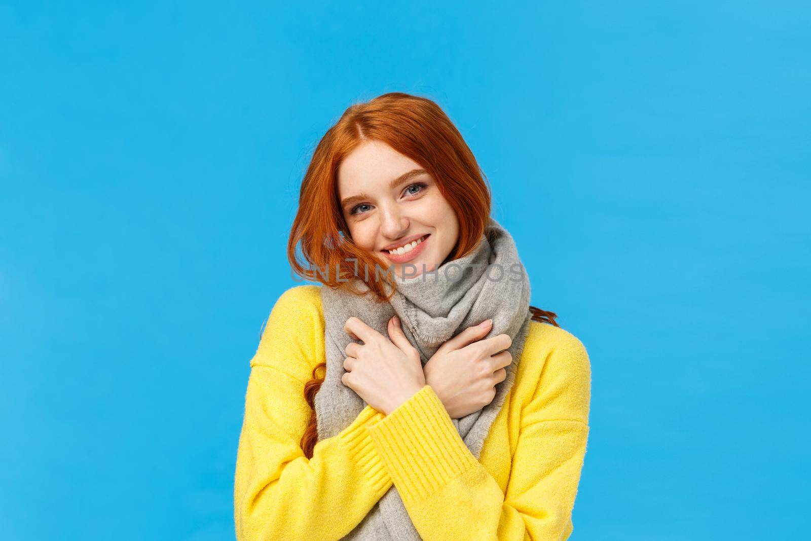 Winter holidays, sales and shopping concept. Waist-up portrait cute and lovely redhead woman wrap neck with soft warm scarf, tilt head smiling happily, embrace herself, standing blue background.