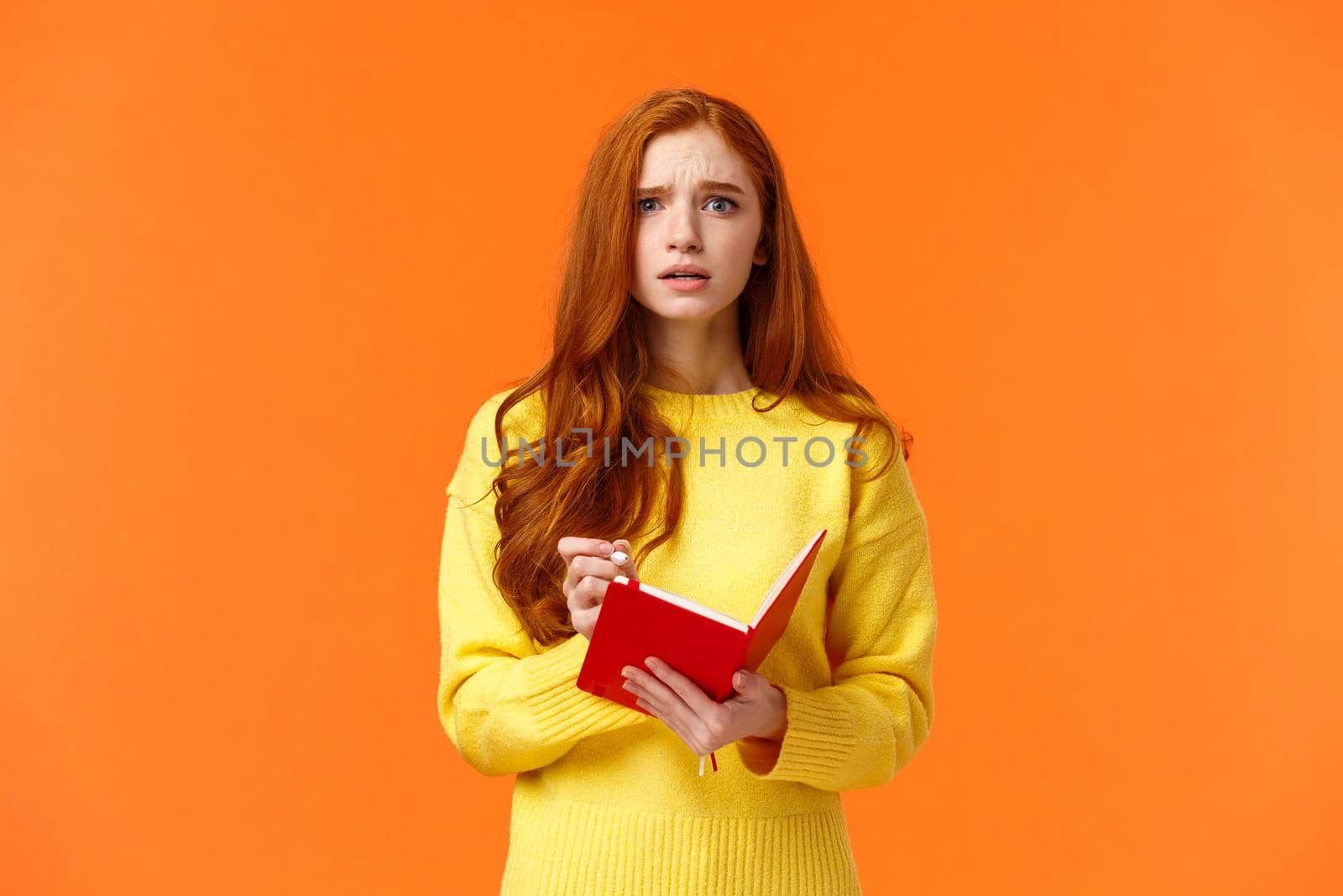 Concerned tensed attractive redhead female student cant write down everything from blackboard after lector, biting lip look troubled and perplexed, holding red notebook and pen, orange background.