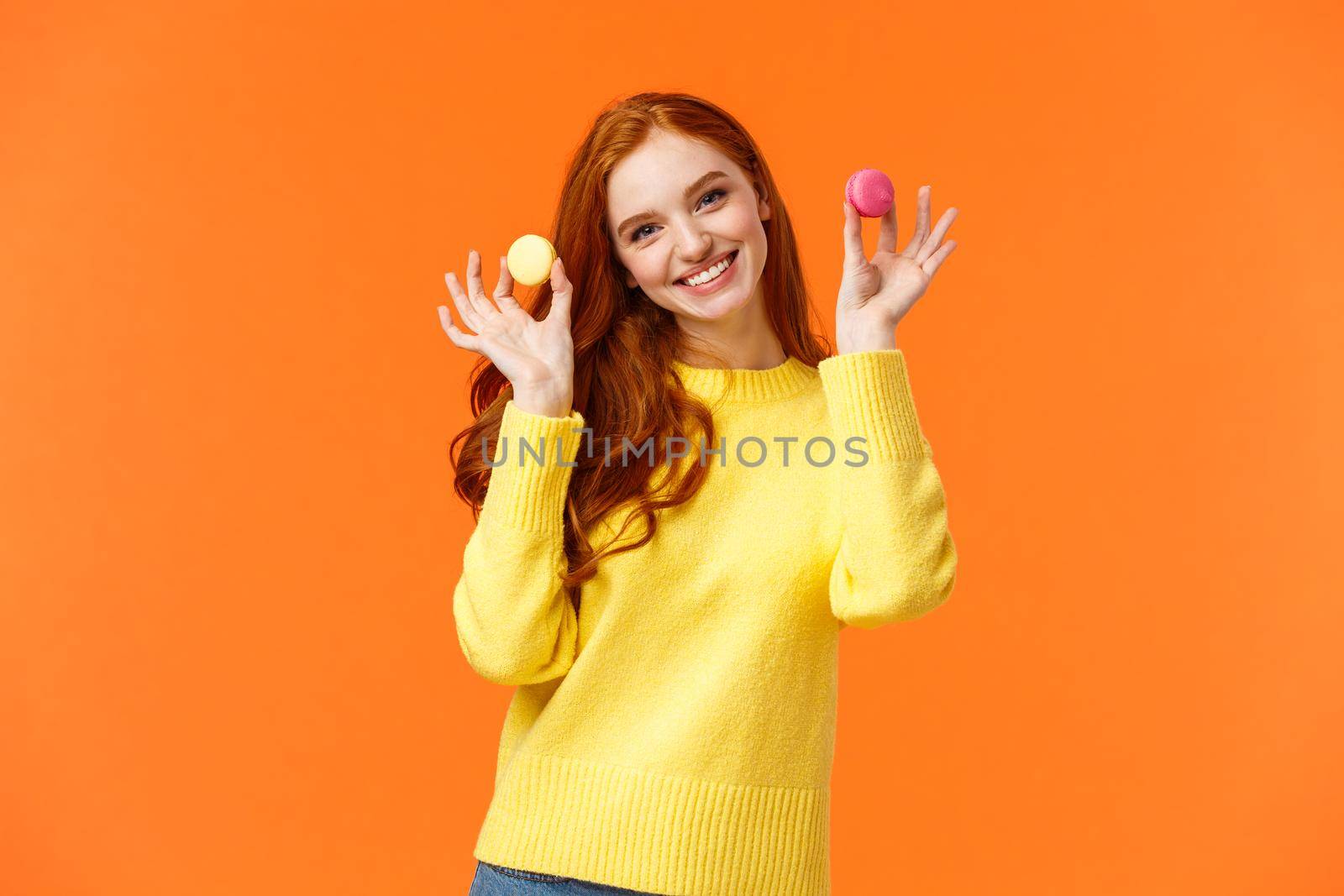 Girlfriend want share dessert, holding two macarons and smiling, suggest bite with lovely grin, tilt head flirty, standing happy orange background, enjoy winter holidays and sweet gifts.