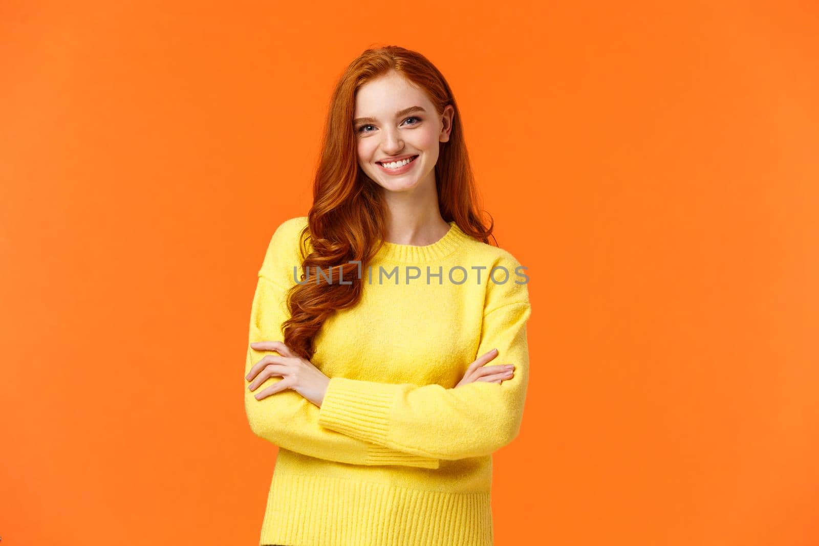 Holidays, people and winter concept. Attractive young smiling girl with red hair in yellow sweater standing in powerful, confident posture, grinning camera, cross arms over chest, orange background.