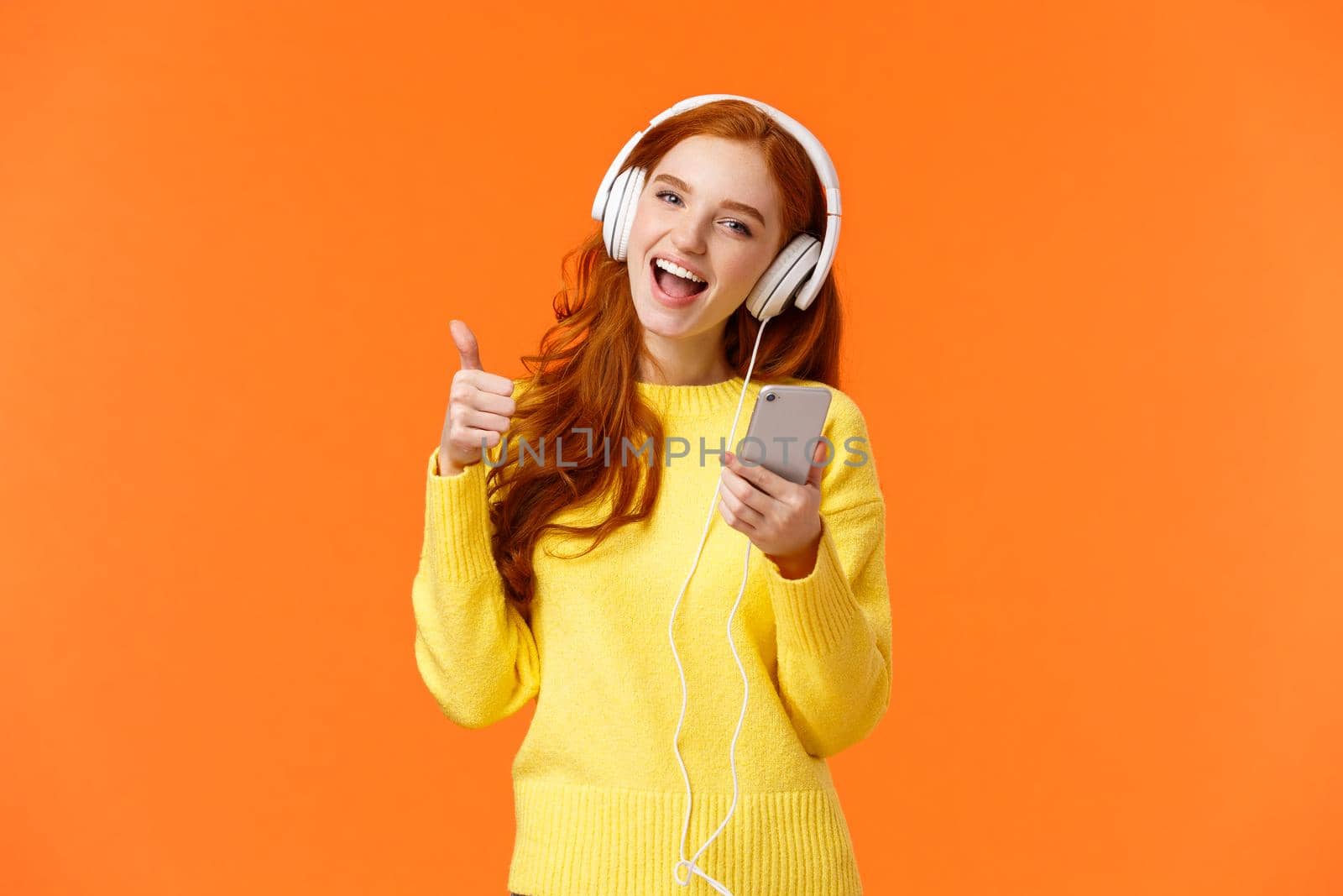 Attractive redhead girl receive christmas gift new cool headphones, listening music in earphones and showing thumb-up, holding smartphone, enjoy music, singing and smiling, orange background.