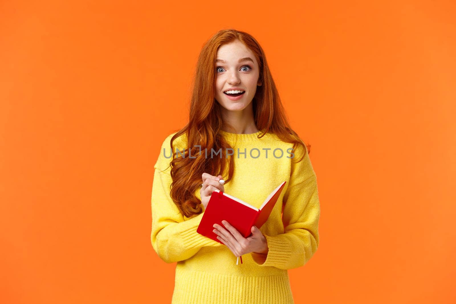 Inspired and upbeat, dreamy fascinated redhead girl writing down interesting lecture, smiling and gazing with admiration or enthusiasm, fill-in notebook schedule, standing orange background by Benzoix