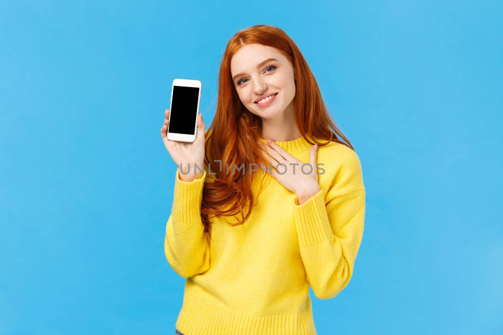 Touched lovely and romantic redhead cute woman bragging with photos from winter holidays, showing smartphone display, touch heart and sighing grateful or pleased, blue background. Copy space