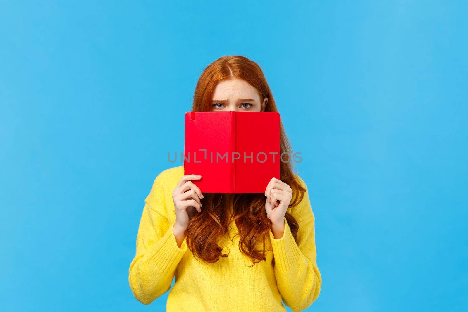 Upset gloomy and uneasy cute pouting redhead girl, frowning dont want speak to you, hiding face behind red notebook, frowning and staring offended, sulking over blue background, bad mood.