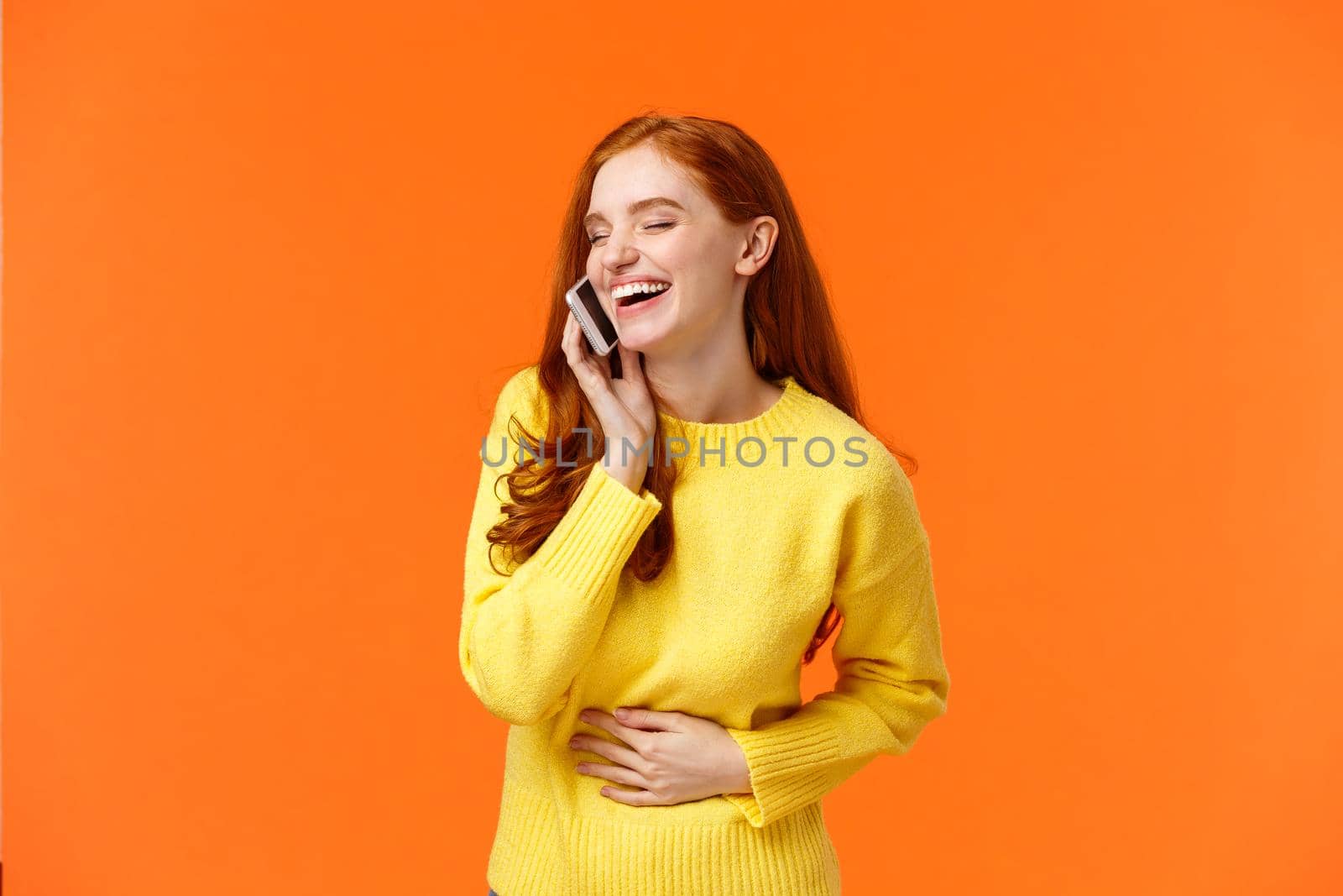 Communication, technology and people concept. Cheerful pretty redhead hipster girl talking on phone and laughing carefree, touching belly close eyes, smartphone near ear, orange background.