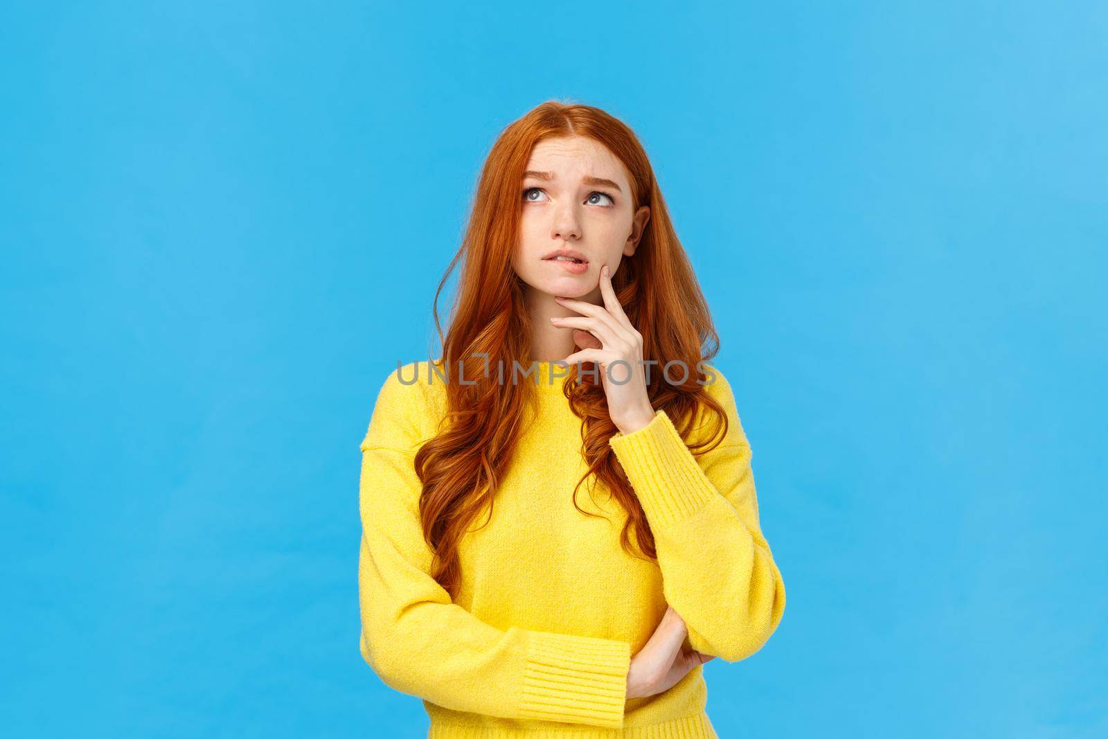 Perplexed, worried and concerned cute redhead student college girl trying choose university, thinking future, looking pensive upper left corner, bite lip and frowning, thoughtful over blue background.
