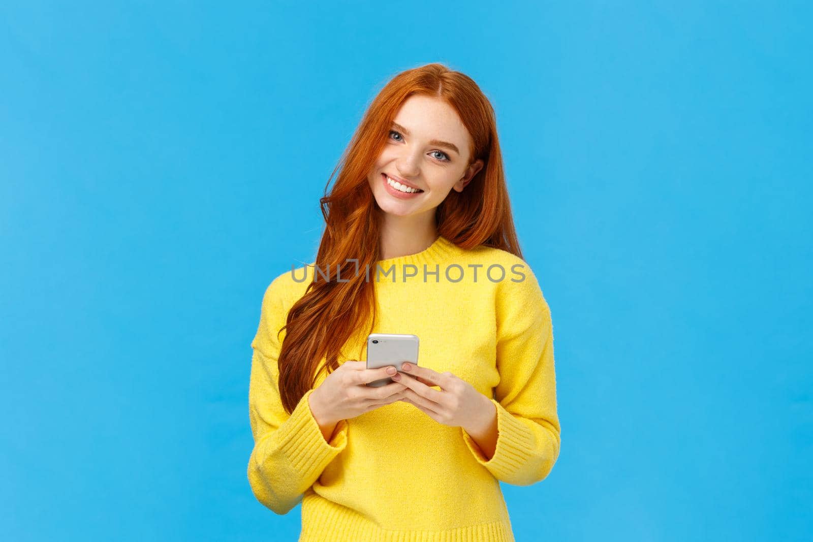 Technology, internet and gadgets concept. Cute redhead woman sending text friend, messaging, having conversation using smartphone app, smiling camera, download useful application, blue background by Benzoix